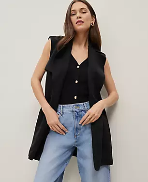 A woman wearing a black Petite Trench Vest with lapels, storm flaps, and a belt, paired with a black top and light blue jeans.