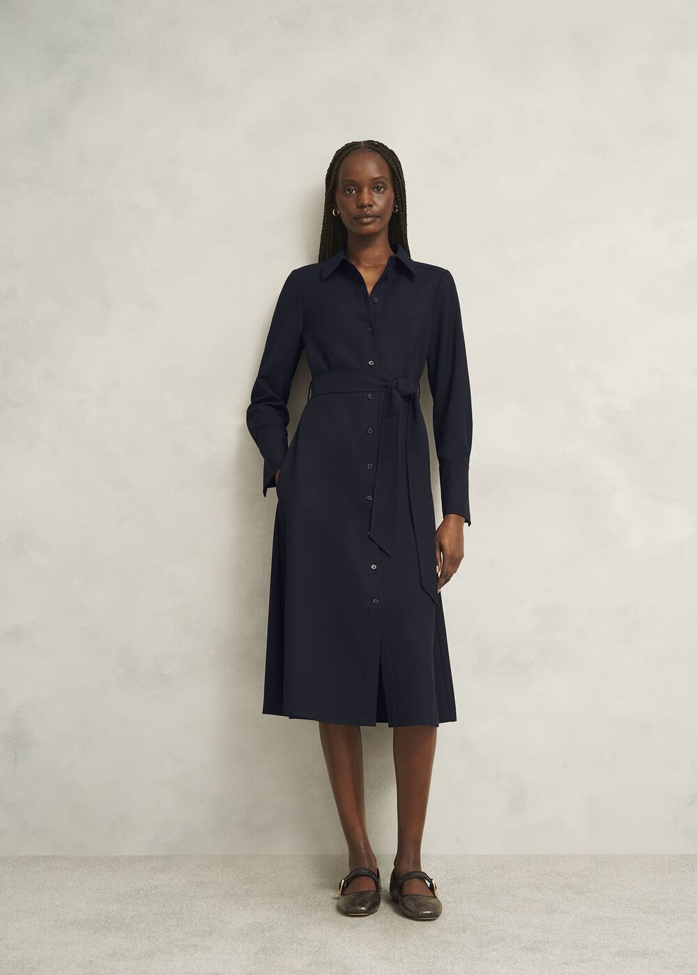 A woman wearing a navy blue shirt dress with a collar, long sleeves, a waist belt, and side pockets, standing against a plain light-colored wall.