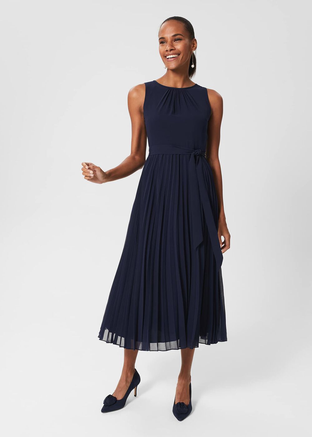 A woman wearing a navy blue sleeveless midi dress with a gathered crew neckline, tie belt at the waist, and pleated skirt, paired with navy high heels.