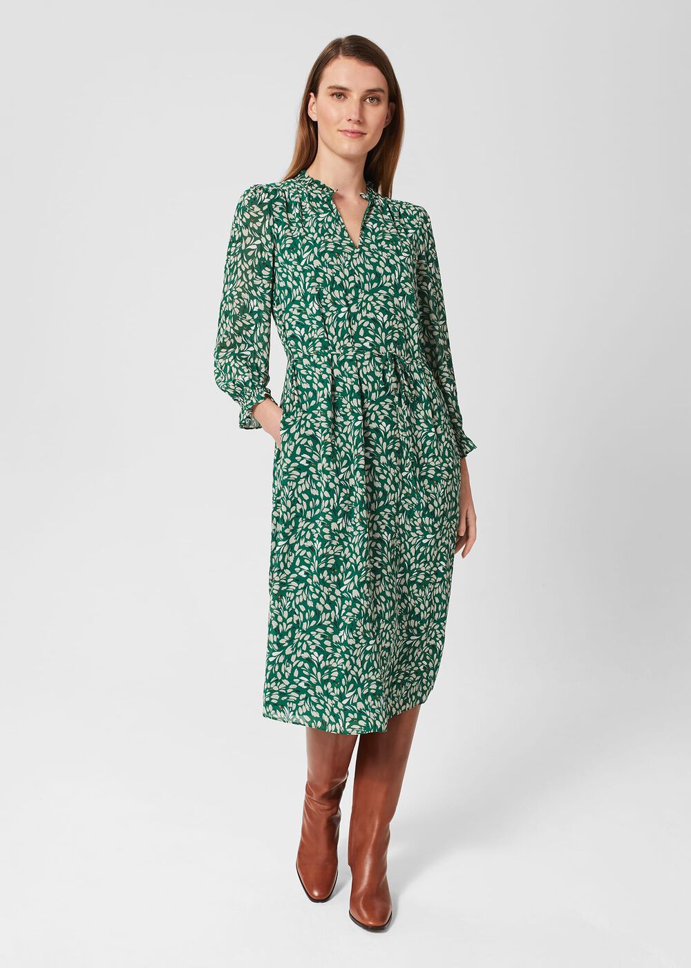 A woman wearing a green floral midi dress with a frill neck and cuffs, tie waist, and side slip pockets.