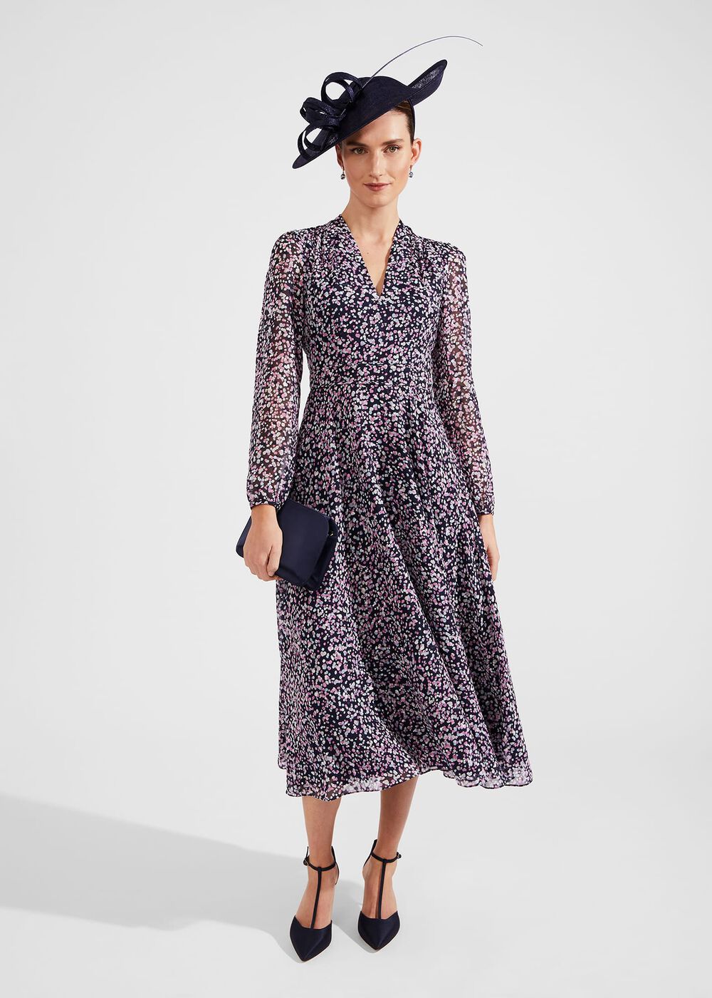 A woman wearing a black midi dress with pink and navy polka dots, long sheer sleeves, a V-neckline, and waist panel detailing, paired with a navy hat and clutch.
