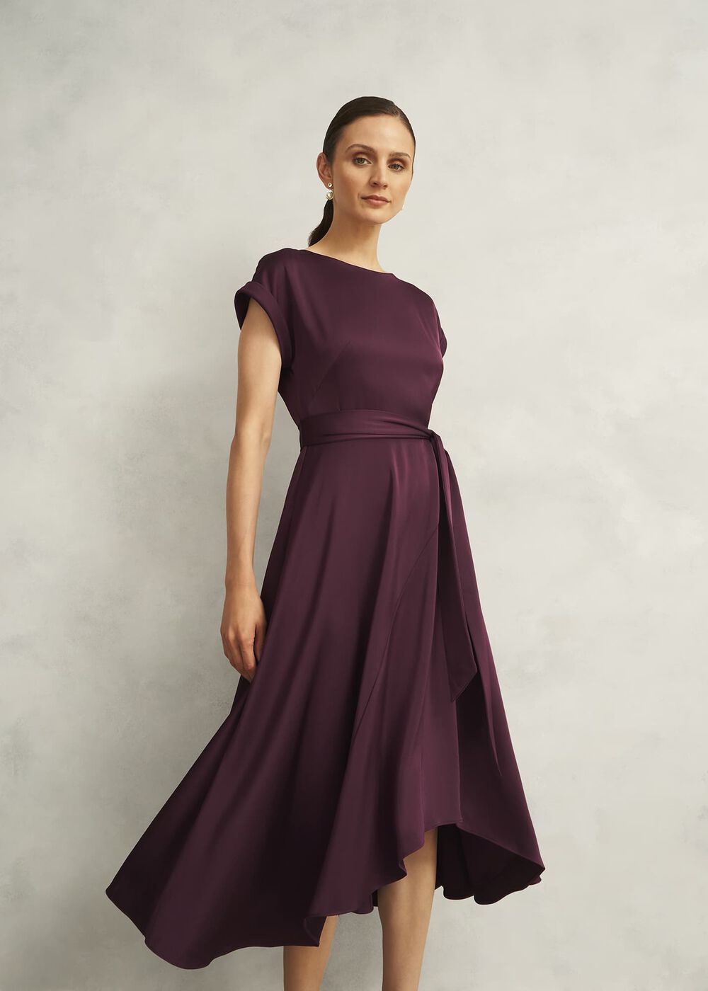 A woman wearing a deep burgundy satin finish dress with a boat neckline, short rolled sleeves, a tie waist, and a curved hem, standing against a plain background.