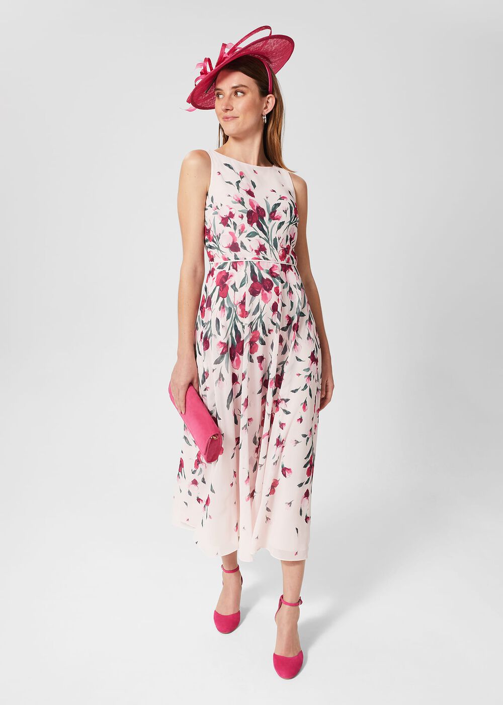 A woman wearing a sleeveless, floral print midi dress with a fit and flare shape, holding a pink clutch, and wearing a large pink hat.