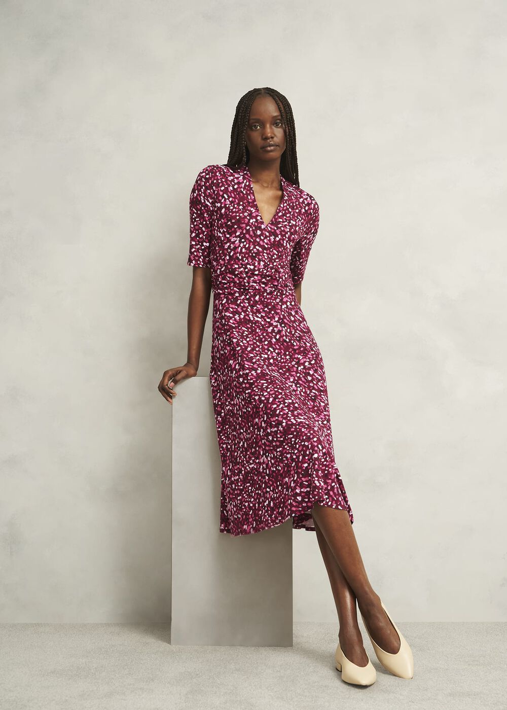 A woman wearing a pink geometric print midi dress with a V neckline and short sleeves, standing against a plain background.