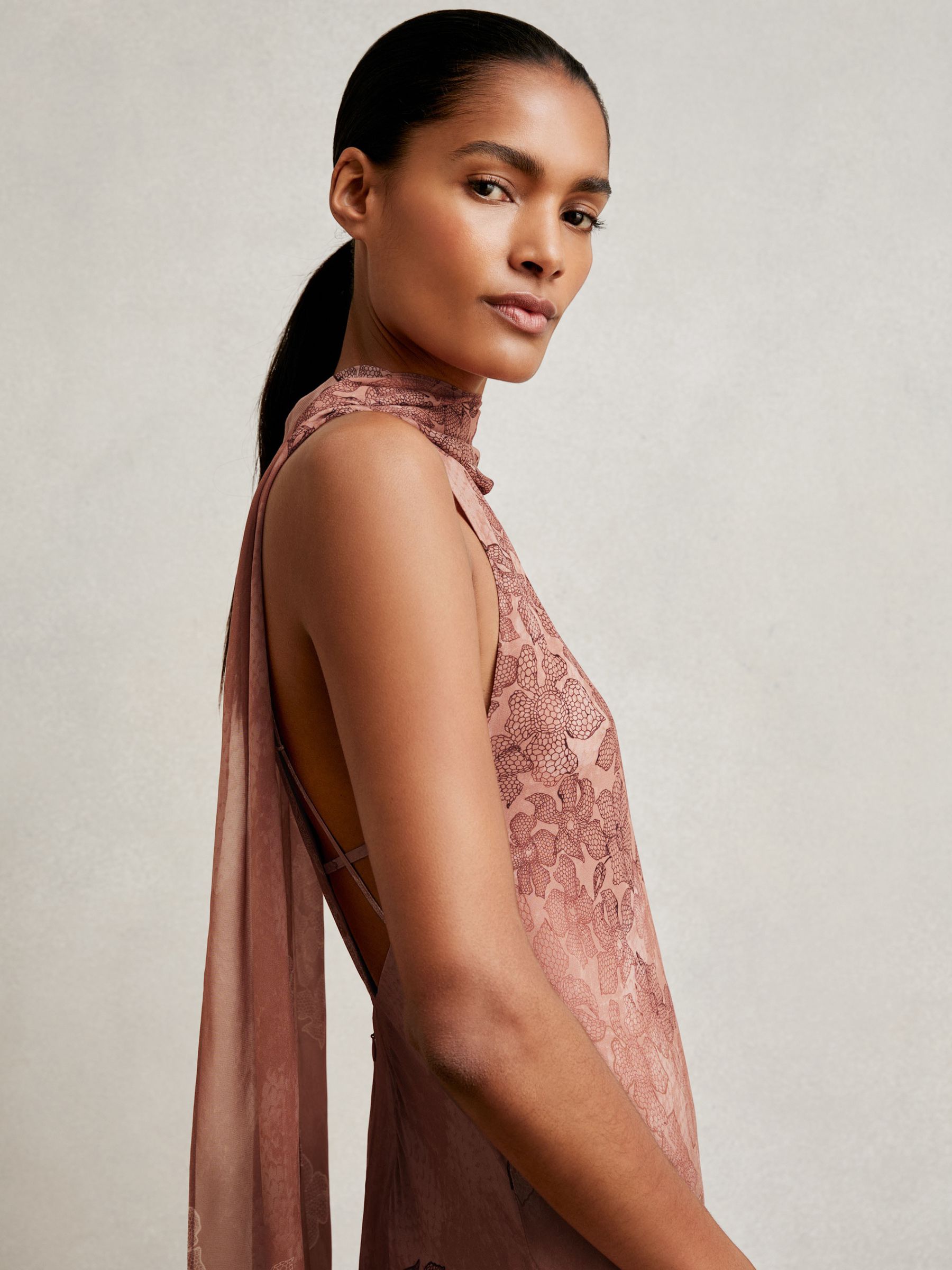 A woman wearing a pale pink chiffon maxi dress with floral print, sleeveless design, soft cowl neckline, and a cape style back with an adjustable strap.