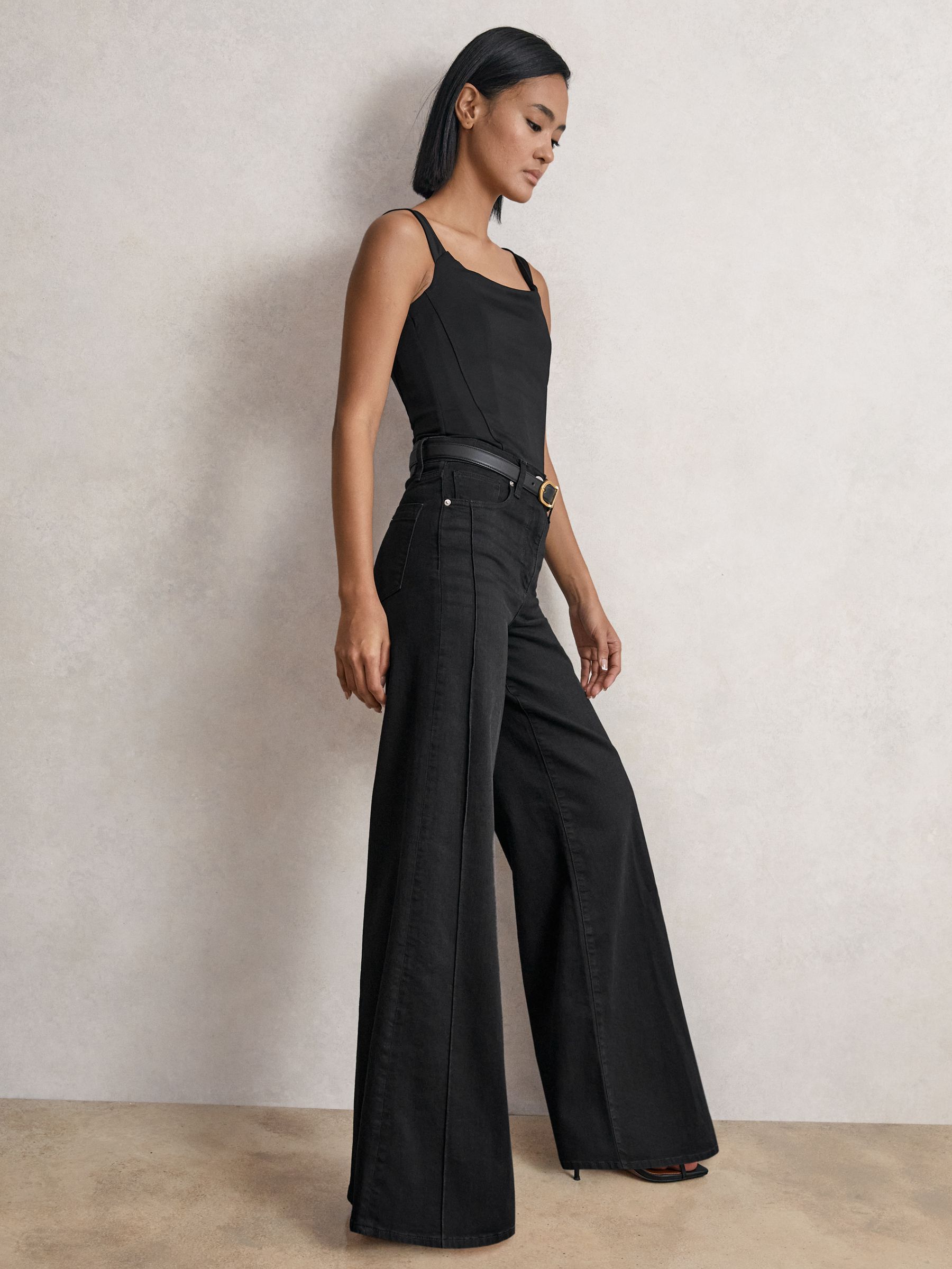 A woman wearing black wide-leg jeans with a high waist, pintucks, and five pockets, paired with a black top.