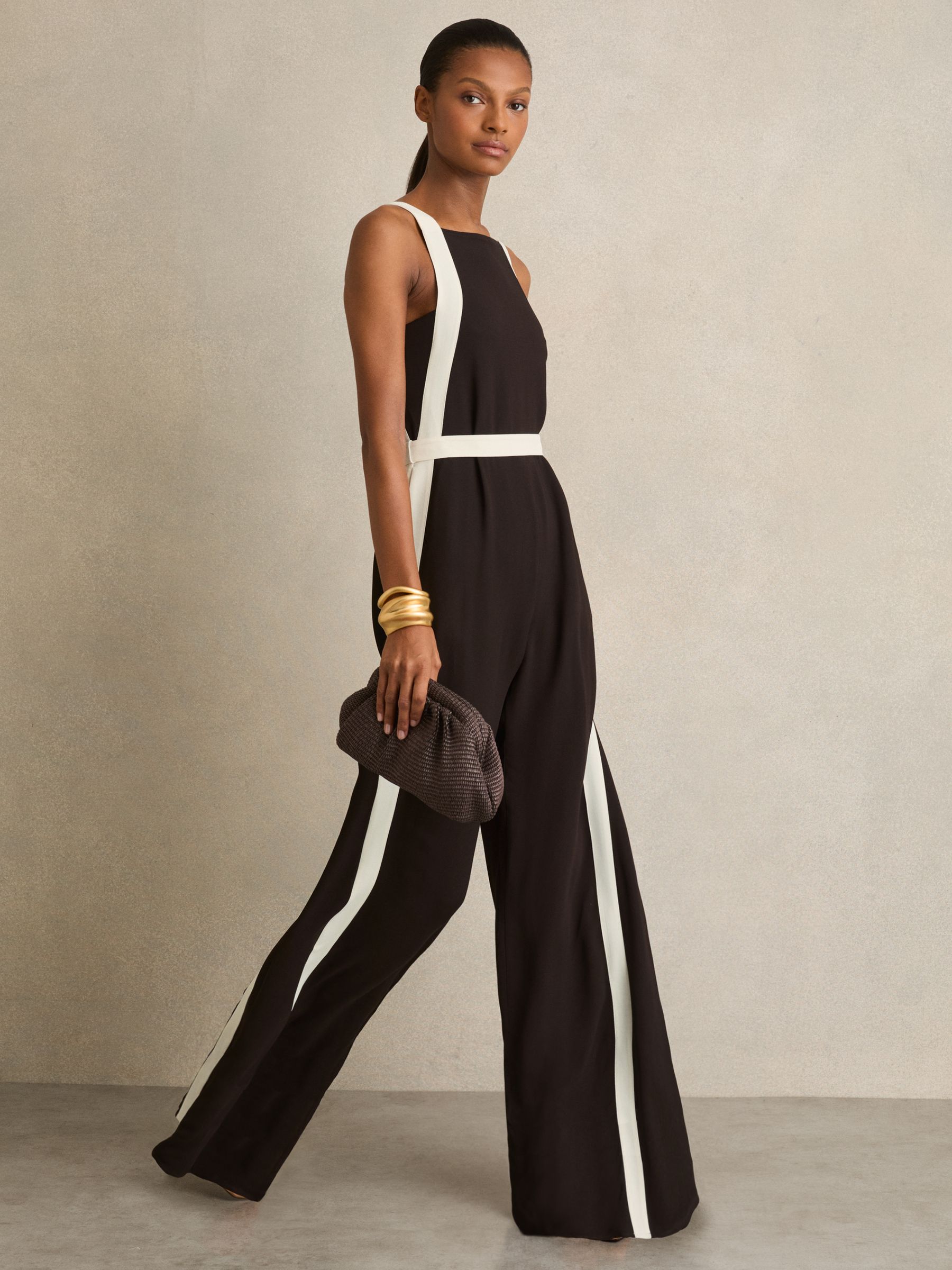 A woman wearing a black jumpsuit with cream panels on the sides, wide-leg silhouette, square neckline, and a self-tie belt, holding a small textured clutch and wearing gold bangles.