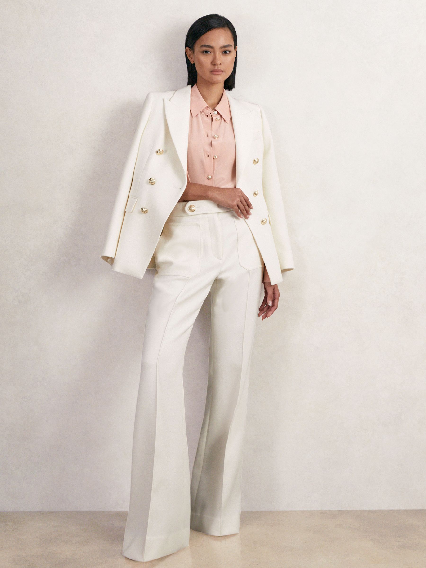 A woman wearing white petite trousers with a high waist, flared legs, front patch pockets, and a matching blazer.