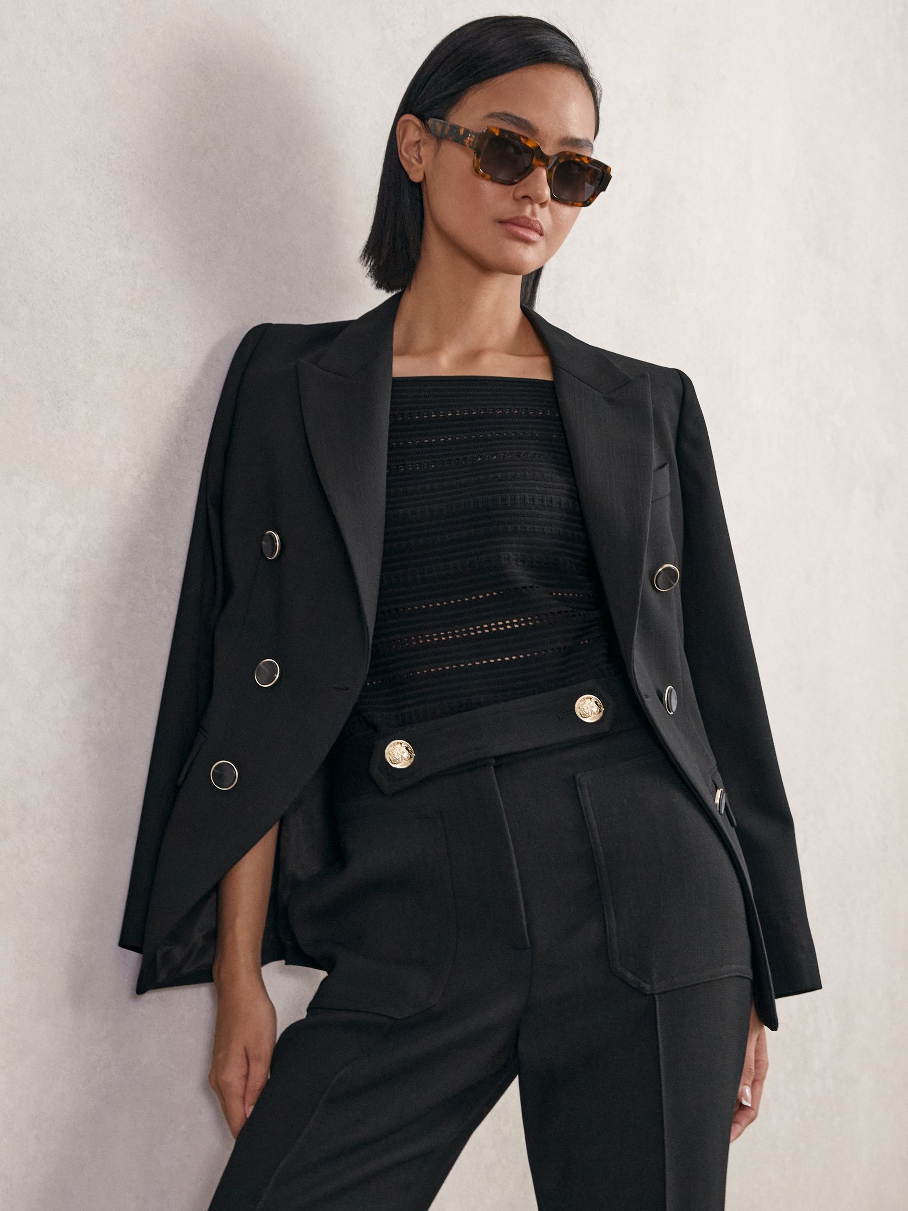 A woman wearing a black double-breasted blazer with peak lapels, padded shoulders, flap pockets, and a back vent, paired with black trousers and sunglasses.