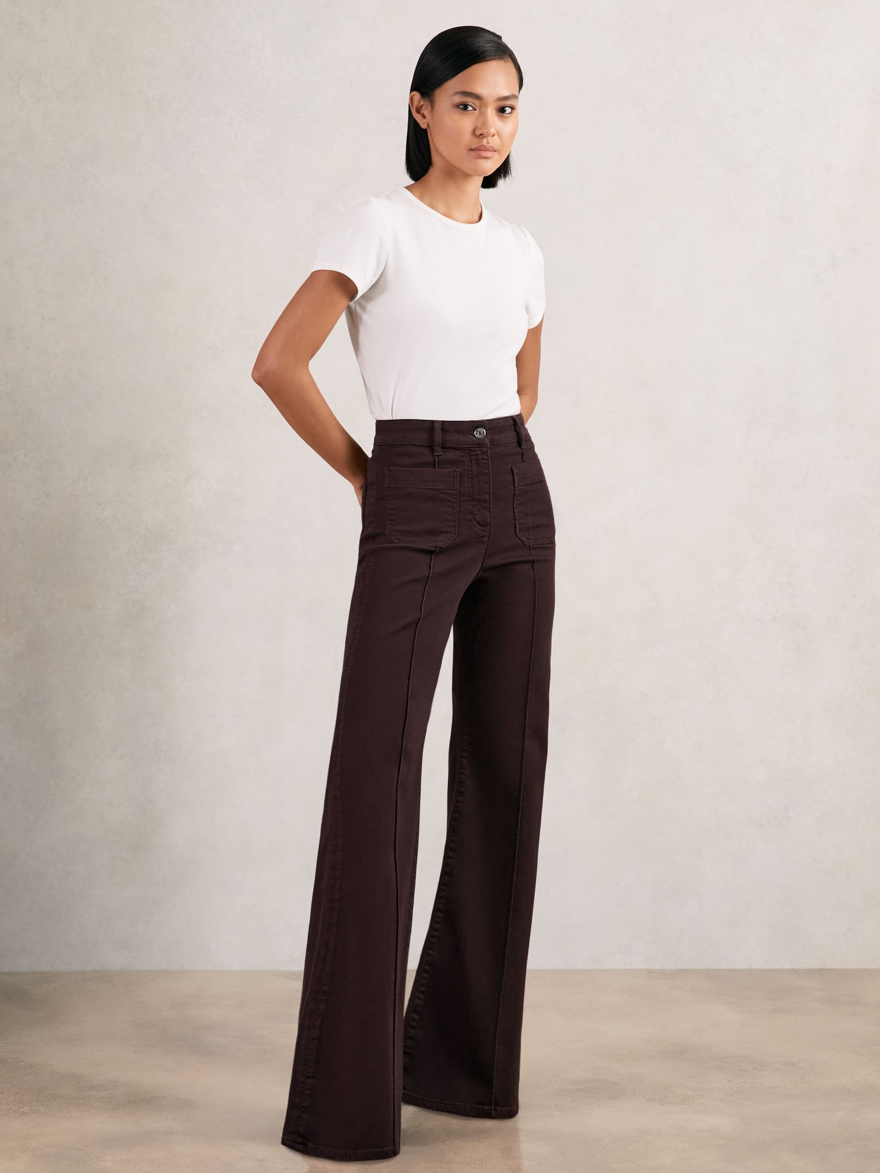 A woman wearing burgundy flared jeans with patch pockets at the front, high-rise waist, and pintuck details, paired with a white t-shirt.