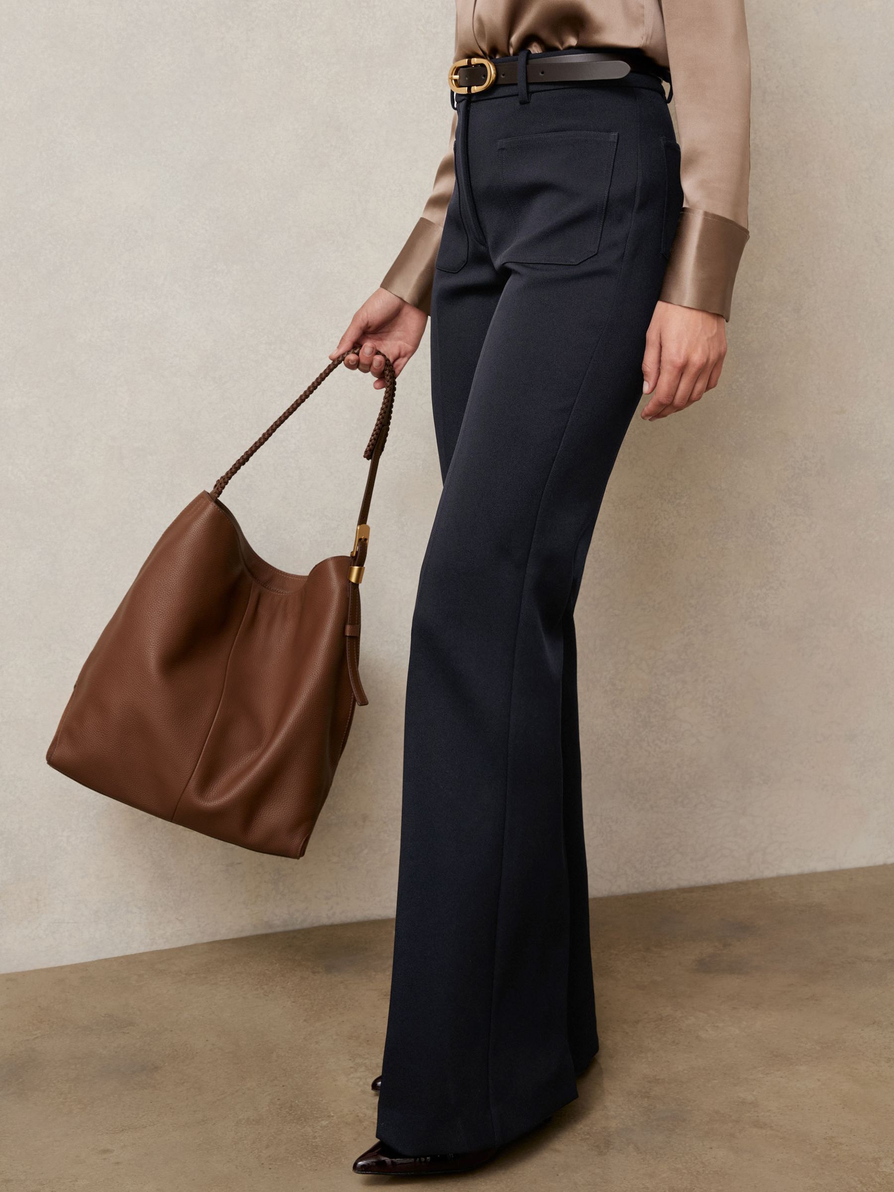 A woman wearing navy flared trousers with patch pockets, a hook and bar fastening, and a zip, paired with a beige satin blouse and holding a brown handbag.