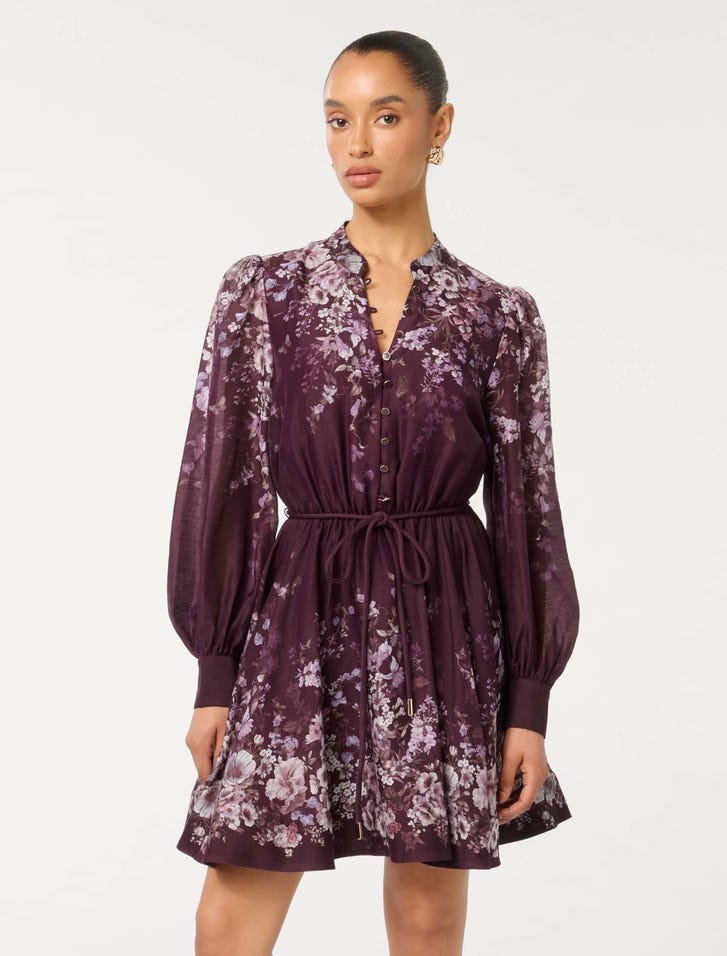 A woman wearing a deep purple floral mini dress with long sleeves, a button front, and a waist tie.