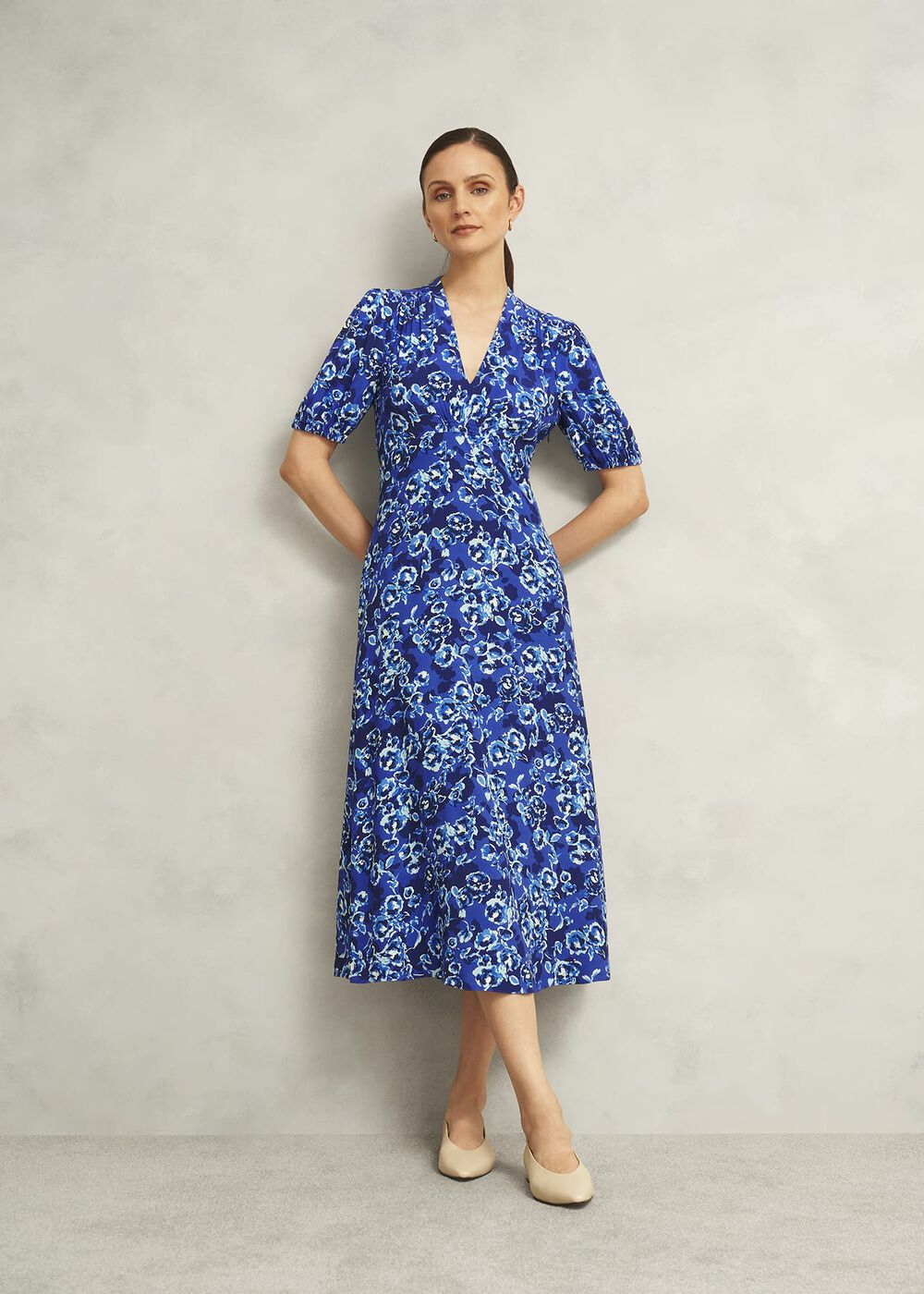 A woman wearing a blue floral print dress with short elasticated sleeves, V-neckline, and side pockets, standing against a plain background.