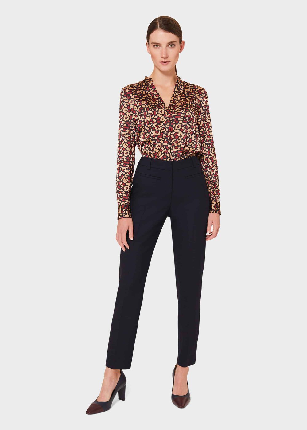 A woman wearing navy tapered trousers with a high waist, belt loops, and pocket details, paired with a patterned blouse.