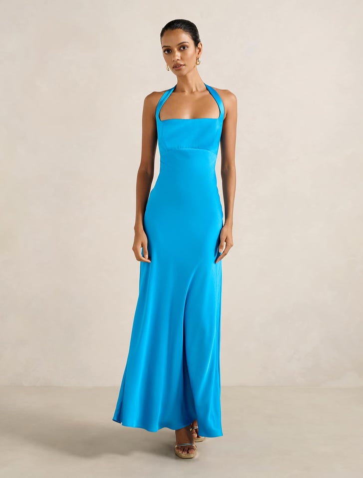 A woman wearing a bright blue satin maxi dress with a halter neckline, sleeveless design, and open back with a metal clasp, standing against a neutral background.