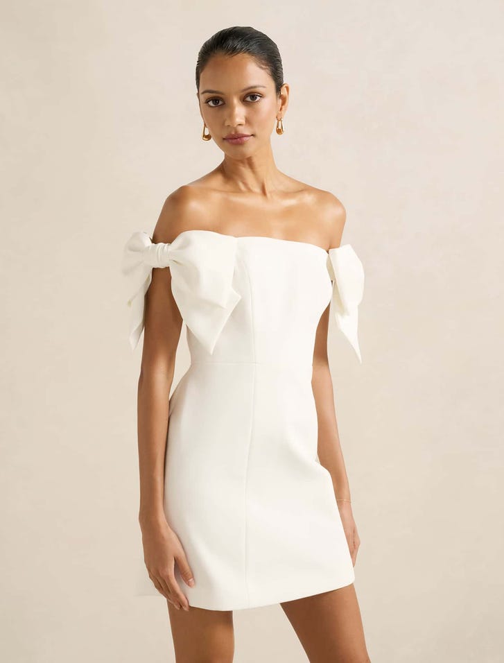 A woman wearing a white off-shoulder mini dress with large bow details on the sleeves, fitted silhouette, and back zip closure.
