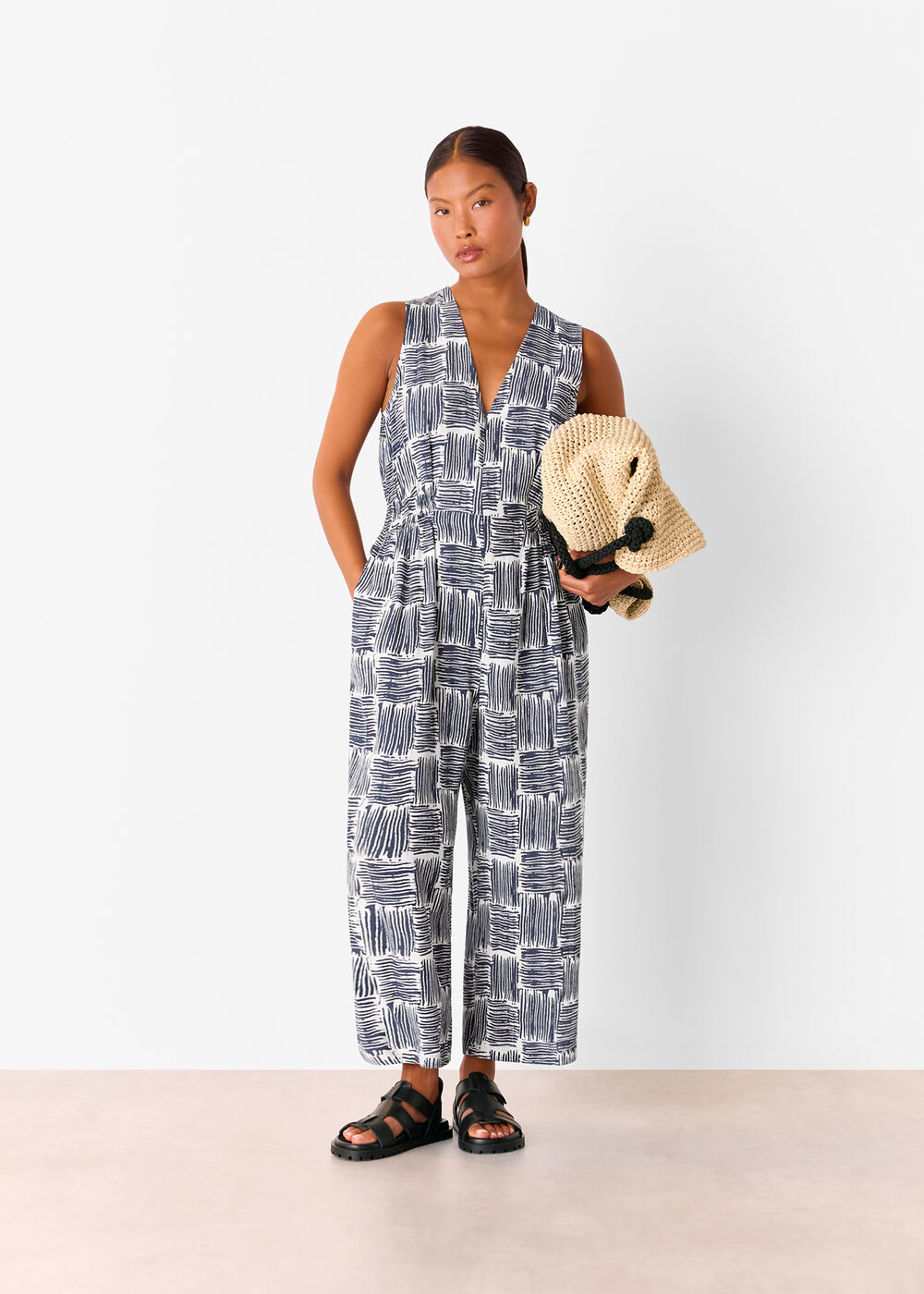 A woman wearing a navy jumpsuit with hatch square pattern, sleeveless, V-neck, and cropped length, holding a beige knit hat with black pom-poms.