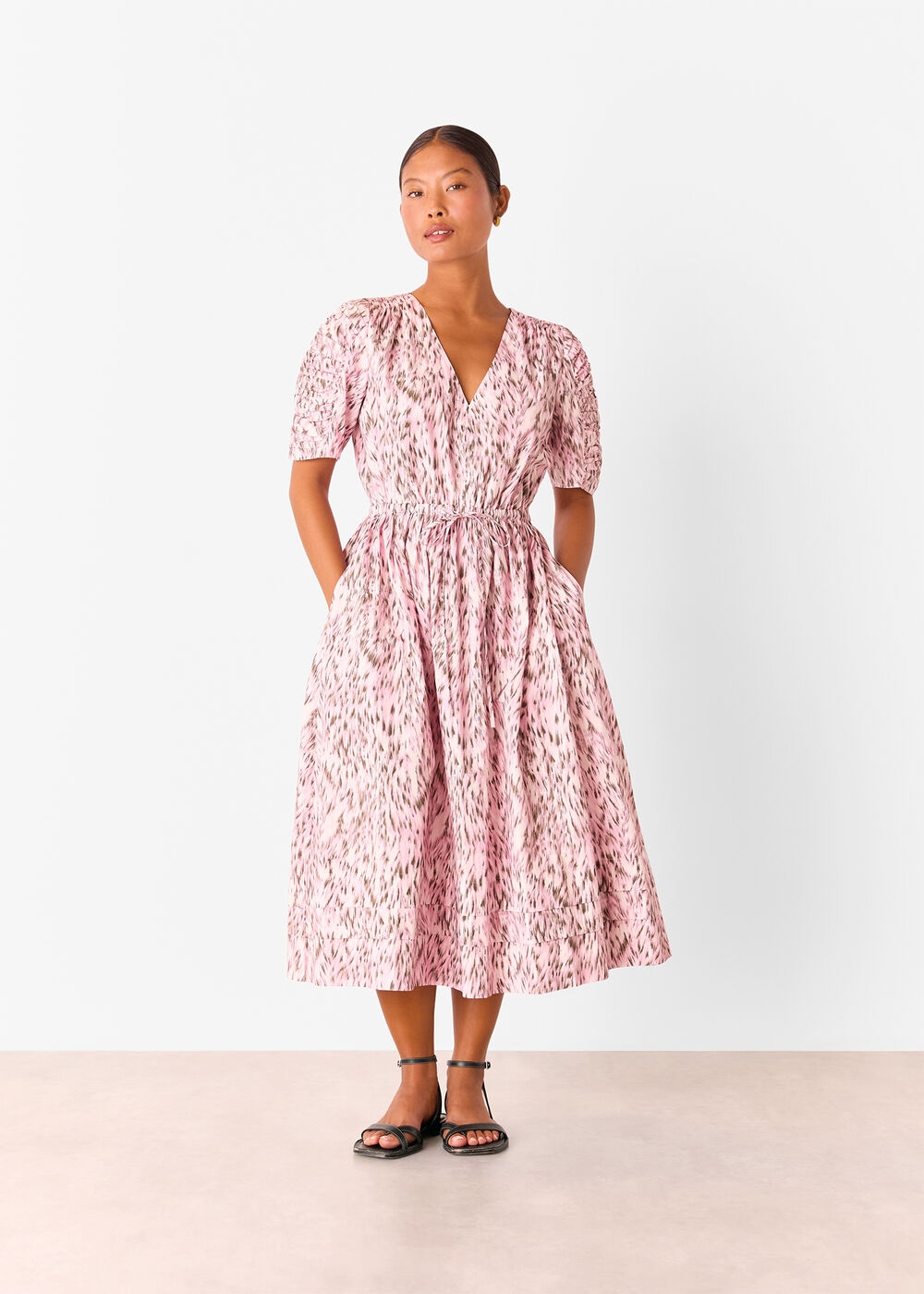 A woman wearing a pink leopard print midi dress with a V-neckline, short puff sleeves, and a waist tie, standing against a plain white background.
