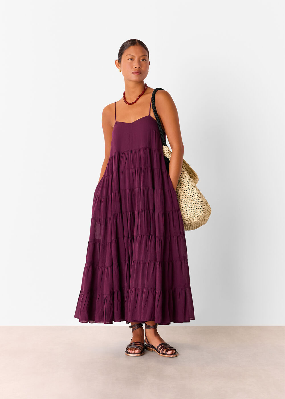 A woman wearing a plum maxi sun dress with thin straps, a curved neckline, and a tiered skirt, accessorized with a red necklace and a woven bag.