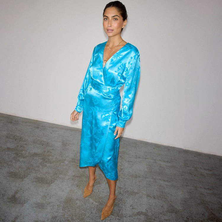 A woman wearing a turquoise wrap dress with a Moroccan-inspired print, long sleeves, and a tie waist, standing against a plain background.
