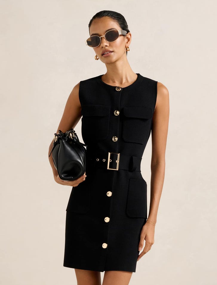 A woman wearing a black sleeveless mini dress with front patch pockets, a belt, and a fitted silhouette, accessorized with sunglasses and a black handbag.