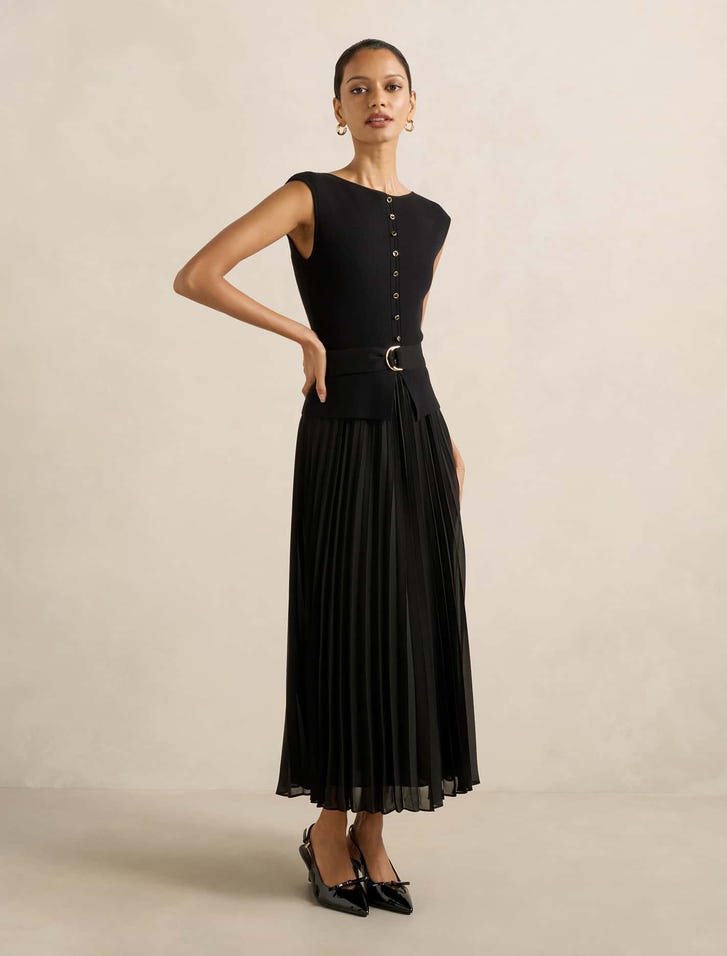 A woman wearing a sleeveless black midi dress with a pleated hem, button-down front, and a belt at the waist, paired with black high heels.
