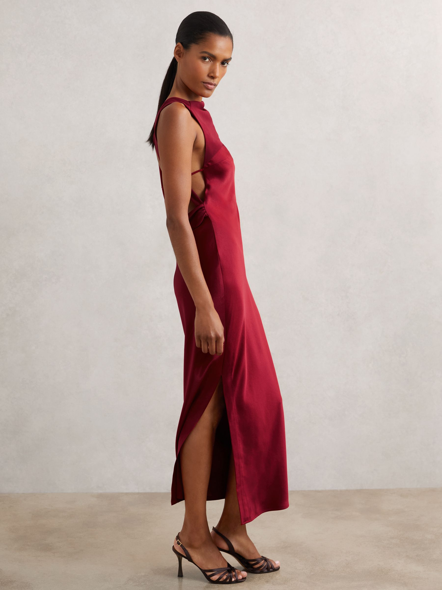 A woman wearing a red satin midi dress with an asymmetric neckline, open back with self-tie straps, side slit, and high heels.