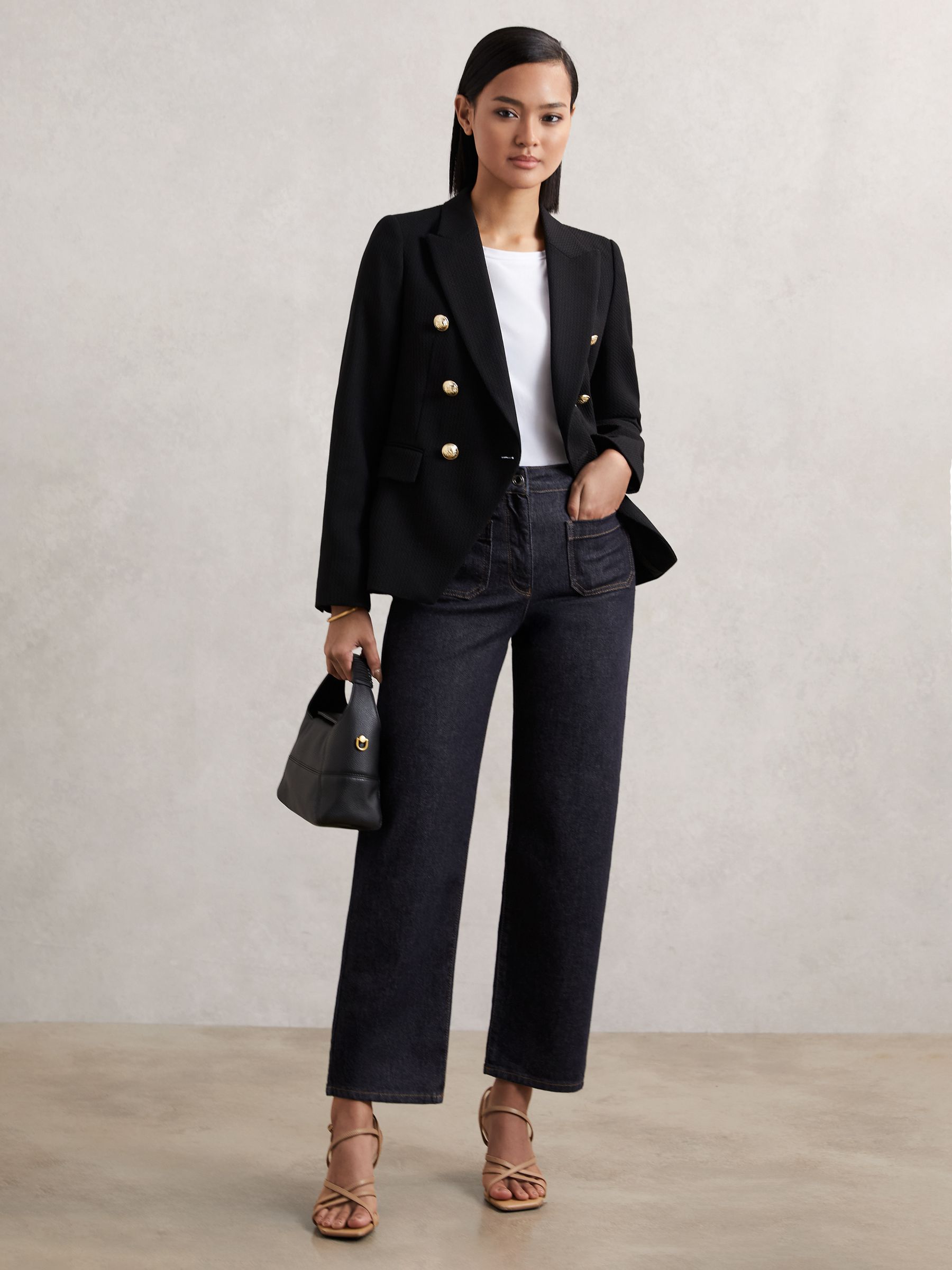 A woman wearing dark blue petite straight leg jeans with patch pockets, a white top, black blazer, and beige heels, holding a black handbag.