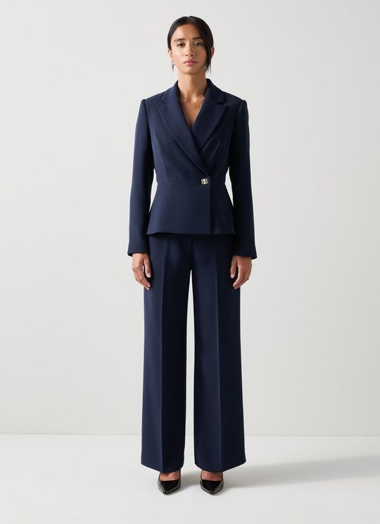A woman wearing a navy tailored jacket with a peplum shape, long sleeves, and a collared neckline, paired with matching navy wide-leg trousers.