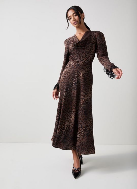 A woman wearing a long leopard print dress with a cowl neckline and lace cuffs, standing against a plain background.