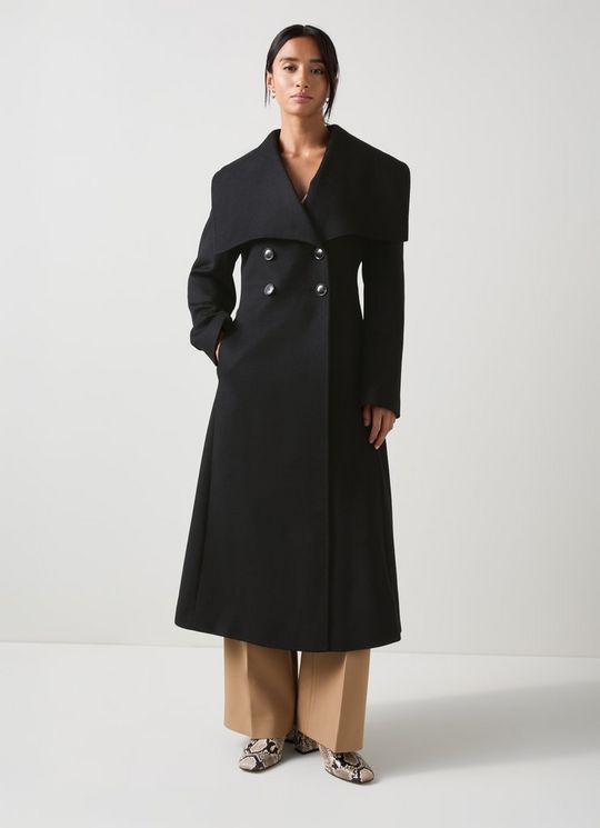 A woman wearing a black fitted coat with a large collar and double-breasted buttons, paired with beige wide-leg trousers and snakeskin shoes.
