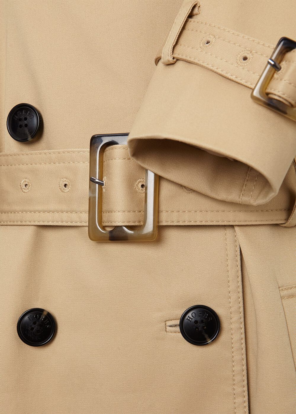 Close-up of a beige women's trench coat with a belt, storm flaps, shoulder epaulettes, and black buttons.