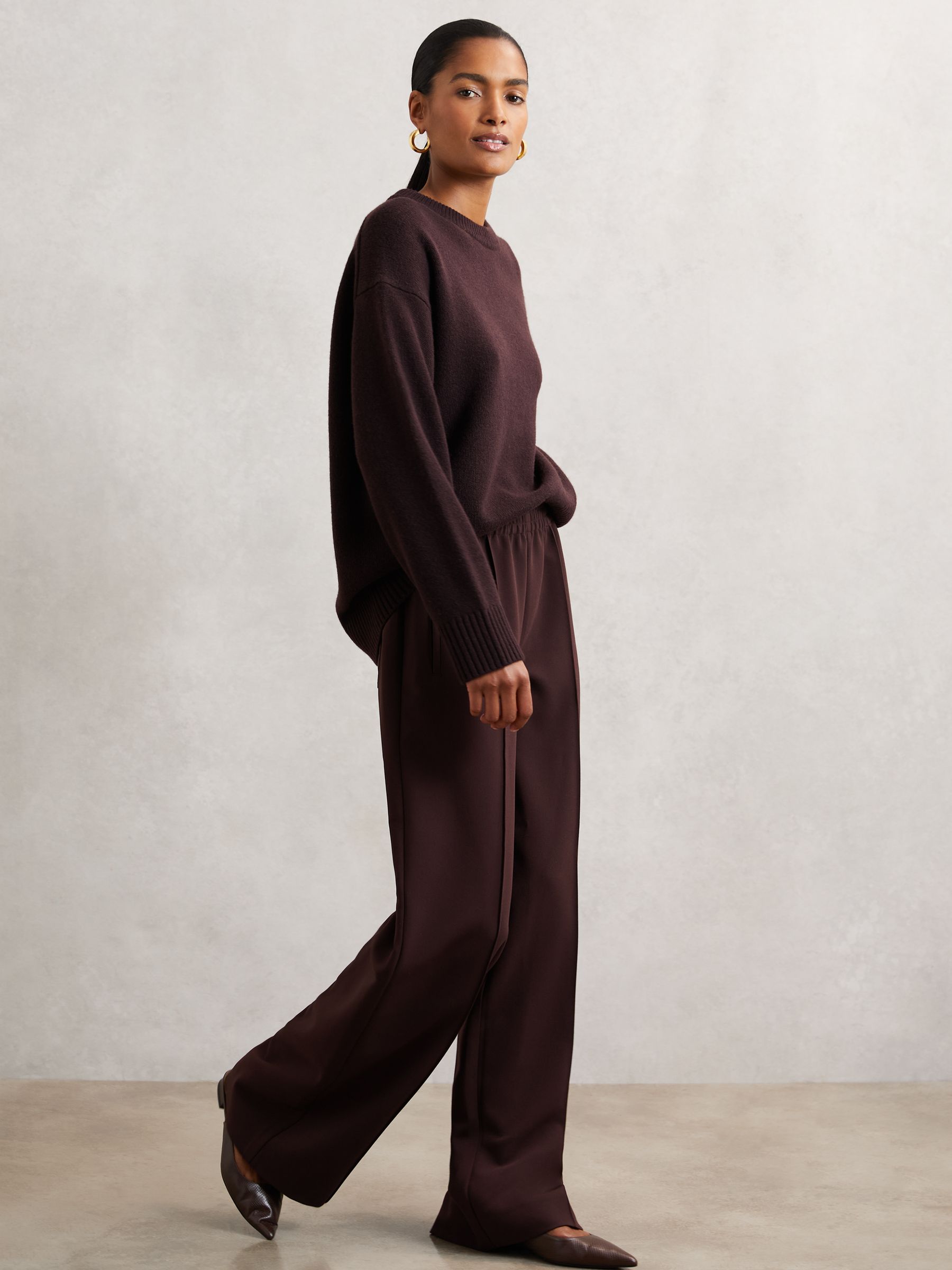 A woman wearing burgundy wide-leg trousers with an elastic waistband, front pintucks, and side slip pockets, paired with a matching burgundy sweater.