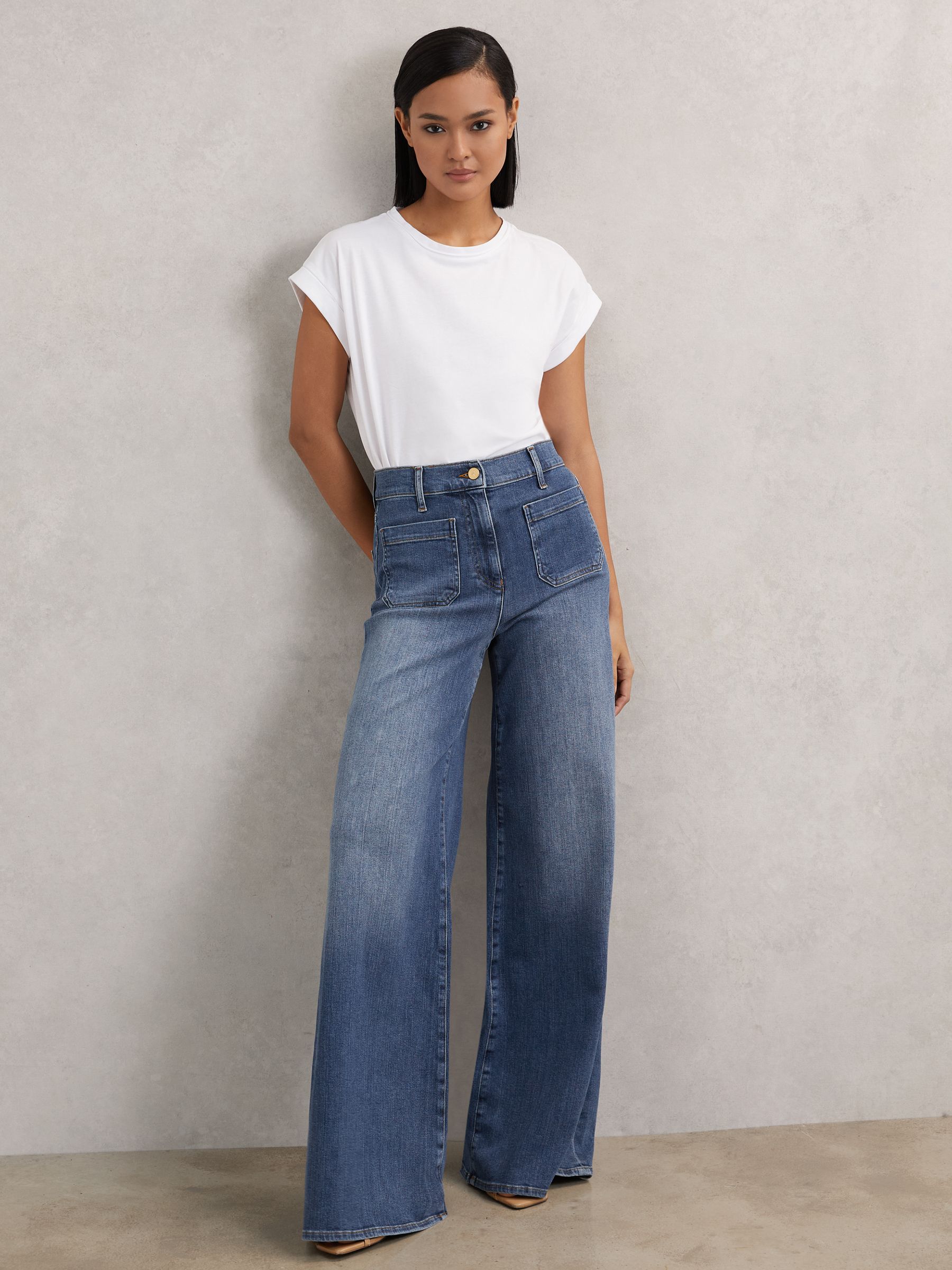 A woman wearing mid blue wide-leg jeans with a high waist, front patch pockets, and a button and zip closure, paired with a white t-shirt.