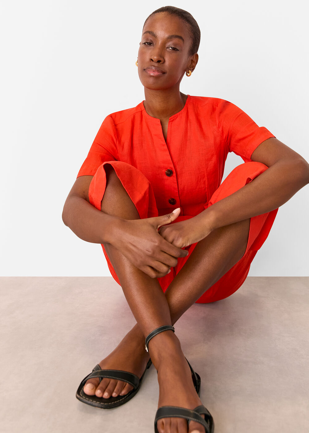 A woman wearing a vibrant red linen jumpsuit with a round neckline, short sleeves, and a belt, sitting with her legs crossed.