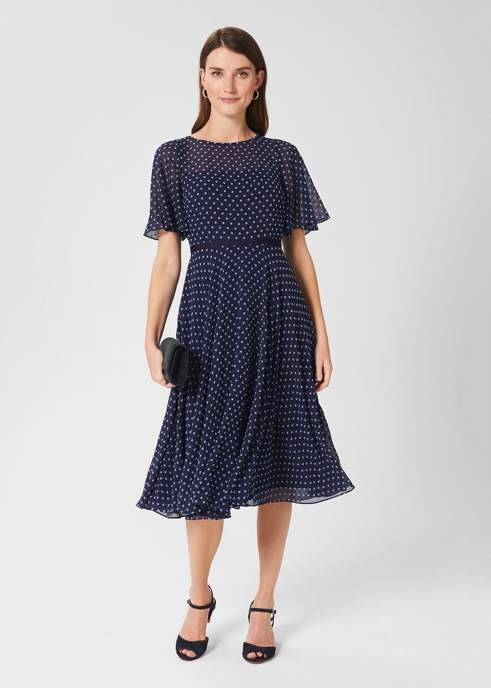 A woman wearing a navy fit and flare dress with white polka dots, fluted sleeves, and a grosgrain waist, holding a black clutch, paired with black high heels.