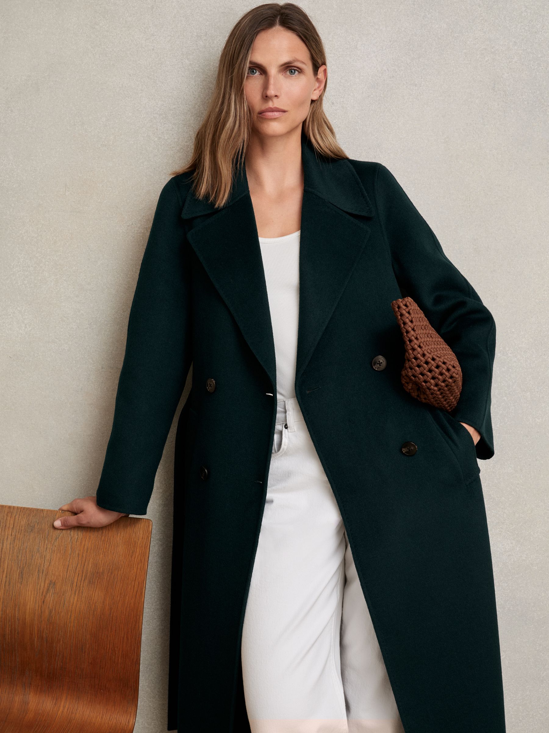A woman wearing a green double-breasted coat with notch lapels, a self-tie belt, and blind seams, made from a wool-blend fabric.