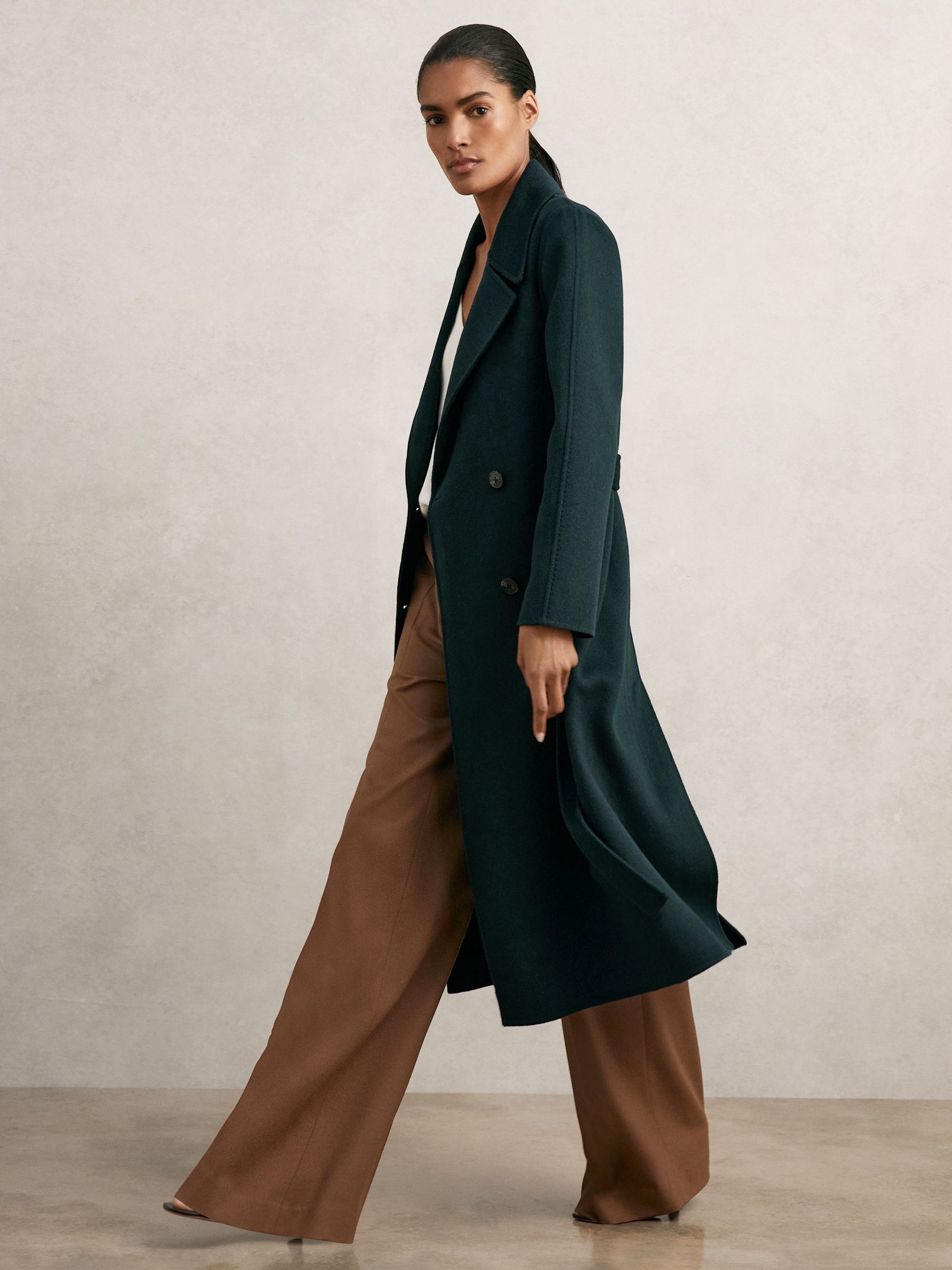 A woman wearing a long green double-breasted coat with notch lapels, blind seams, and a self-tie belt, styled with brown wide-leg trousers.