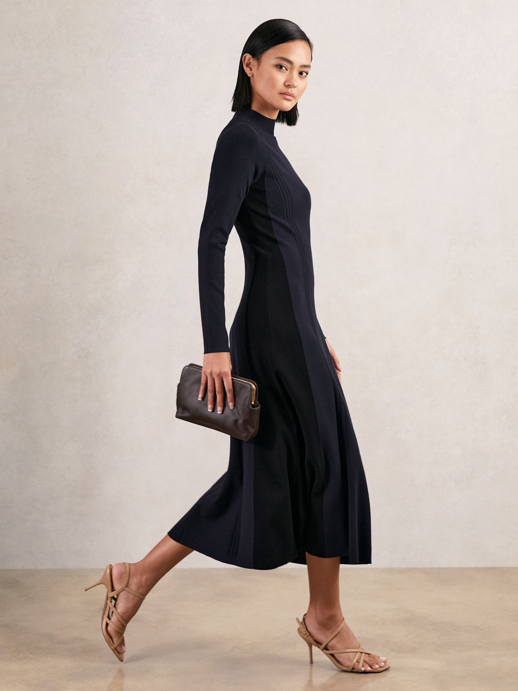A woman wearing a navy and black knitted midi dress with contrast panels, ribbed details, and a fluted skirt, holding a brown clutch and wearing beige high-heeled sandals.