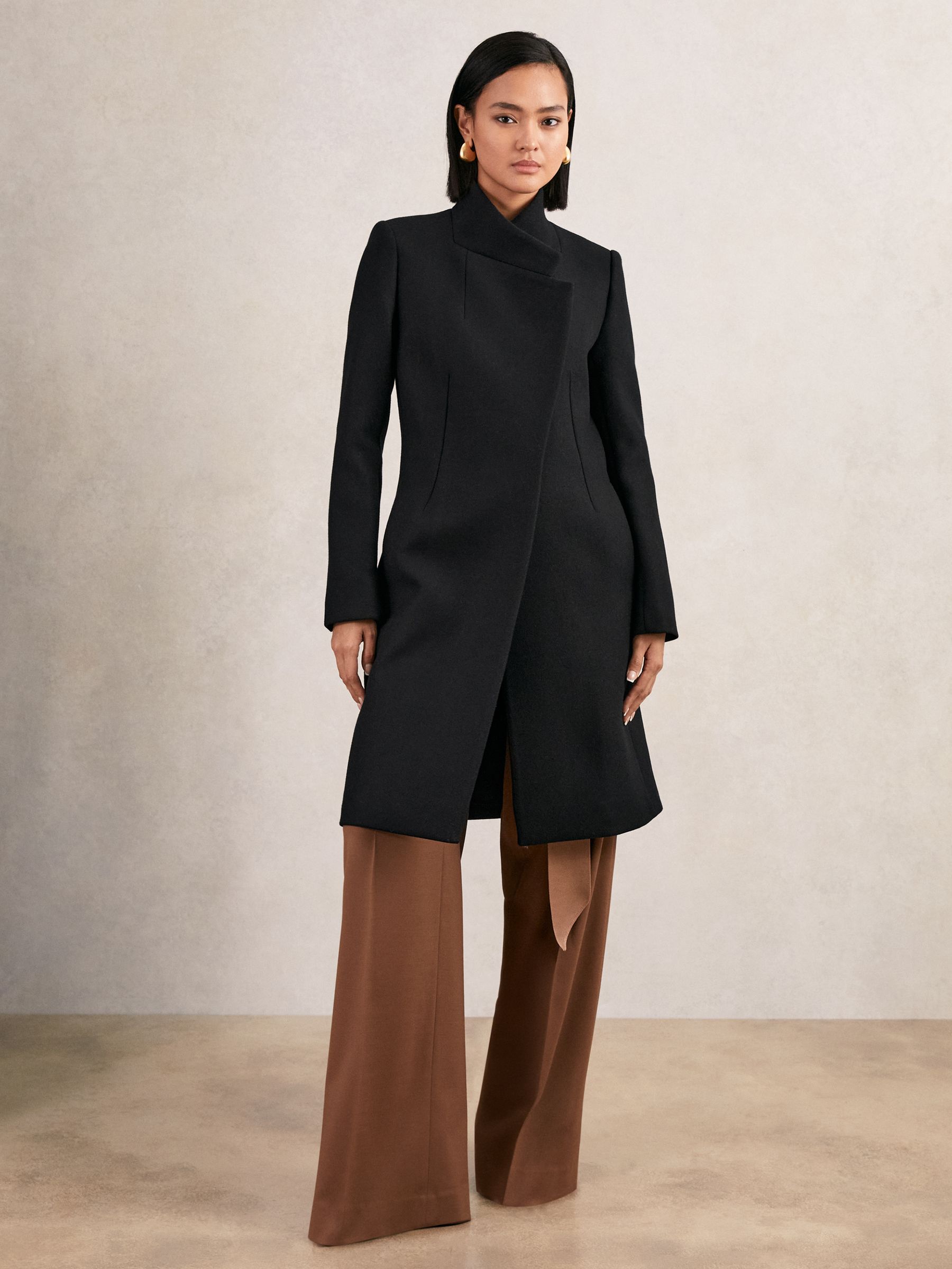 A woman wearing a black, mid-length coat with a wrapover front, standing collar, and concealed buttons.