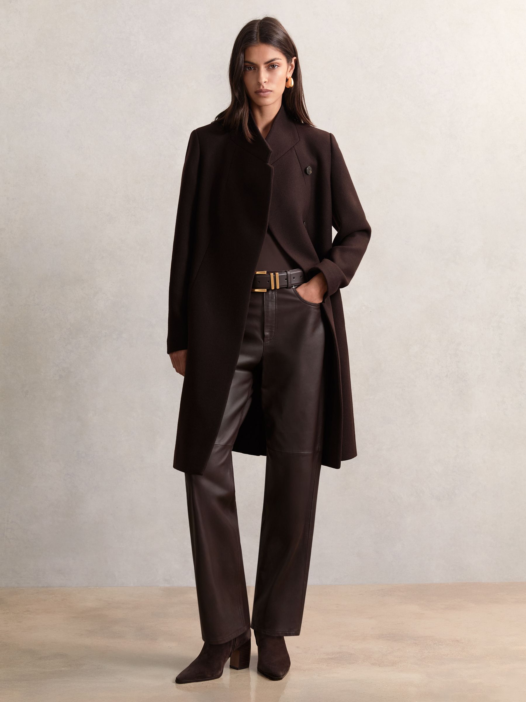 A woman wearing a chocolate brown wool-blend wrapover mid-length coat with a standing collar and concealed buttons, styled with black trousers and boots.
