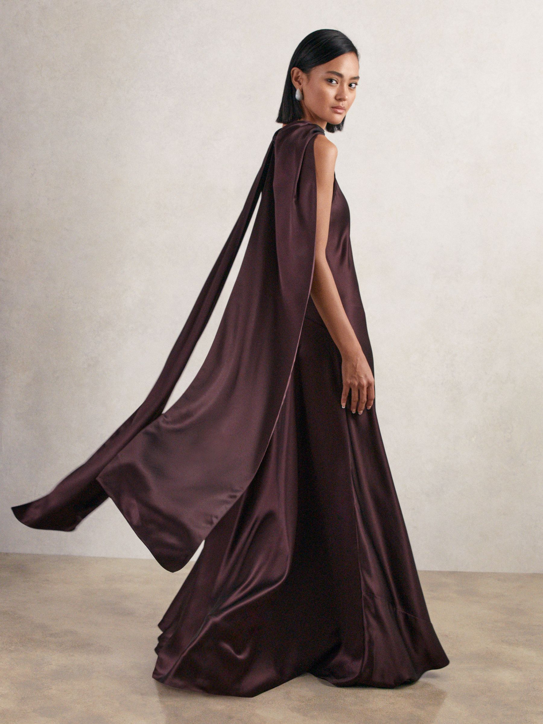 A woman wearing a burgundy satin maxi dress with a cape, sleeveless, with a draped neckline and a concealed zipper.