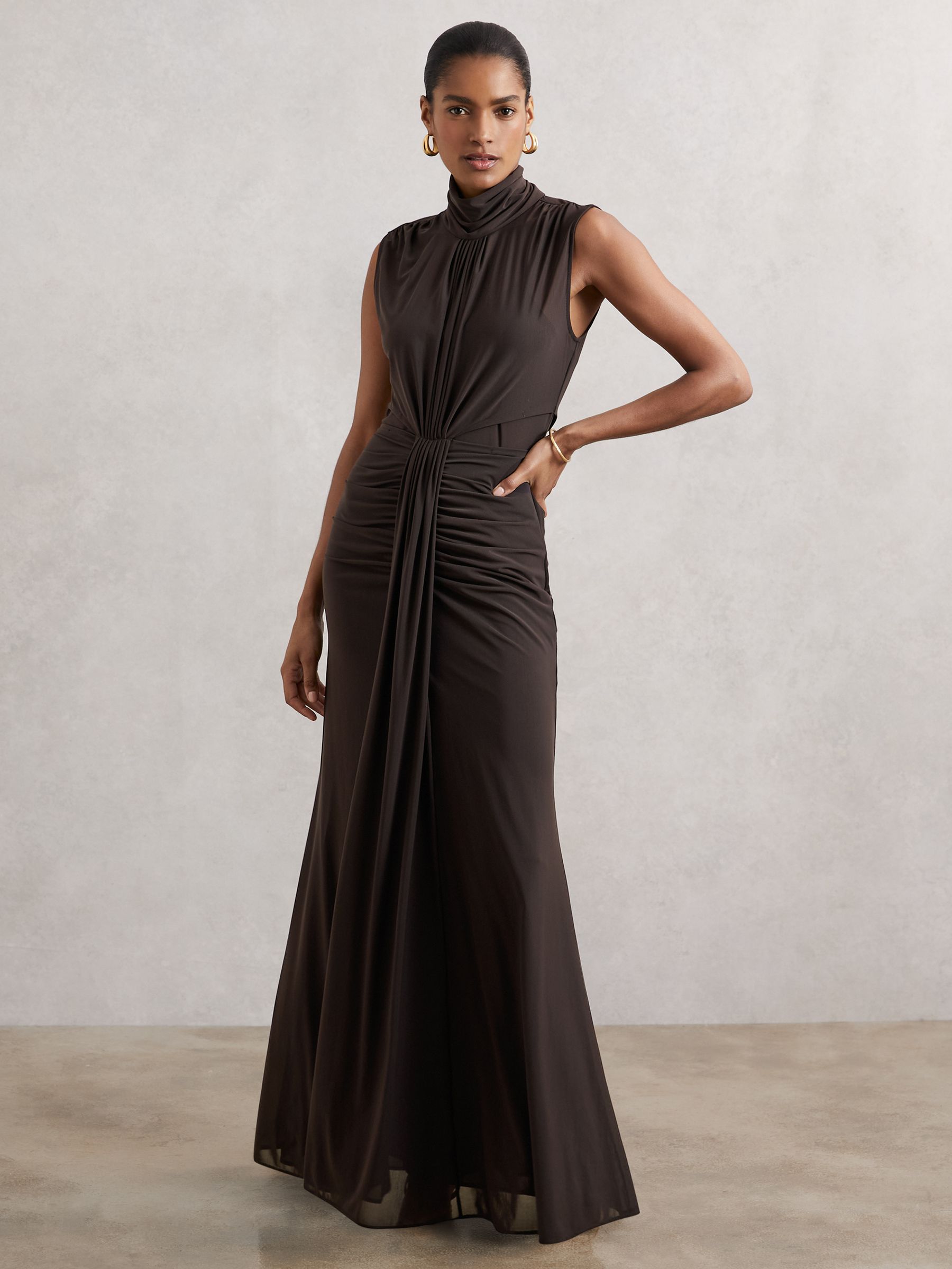 A woman wearing a sleeveless, high-neck, sheer maxi dress in a deep purple grape color with ruched detailing and a flowing silhouette.