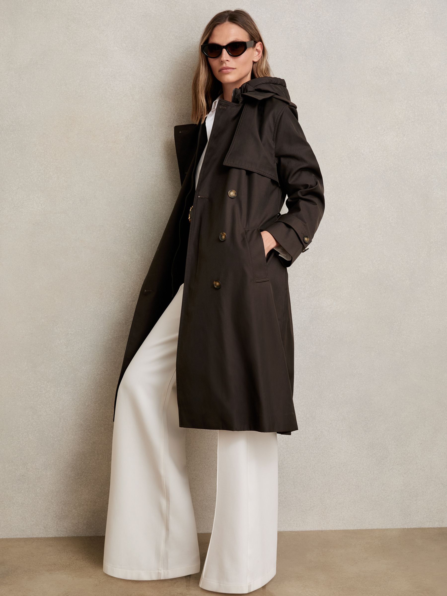 A woman wearing a chocolate brown hooded trench coat with a layered design, quilted hood insert, and gold buttons, paired with white wide-leg pants and sunglasses.