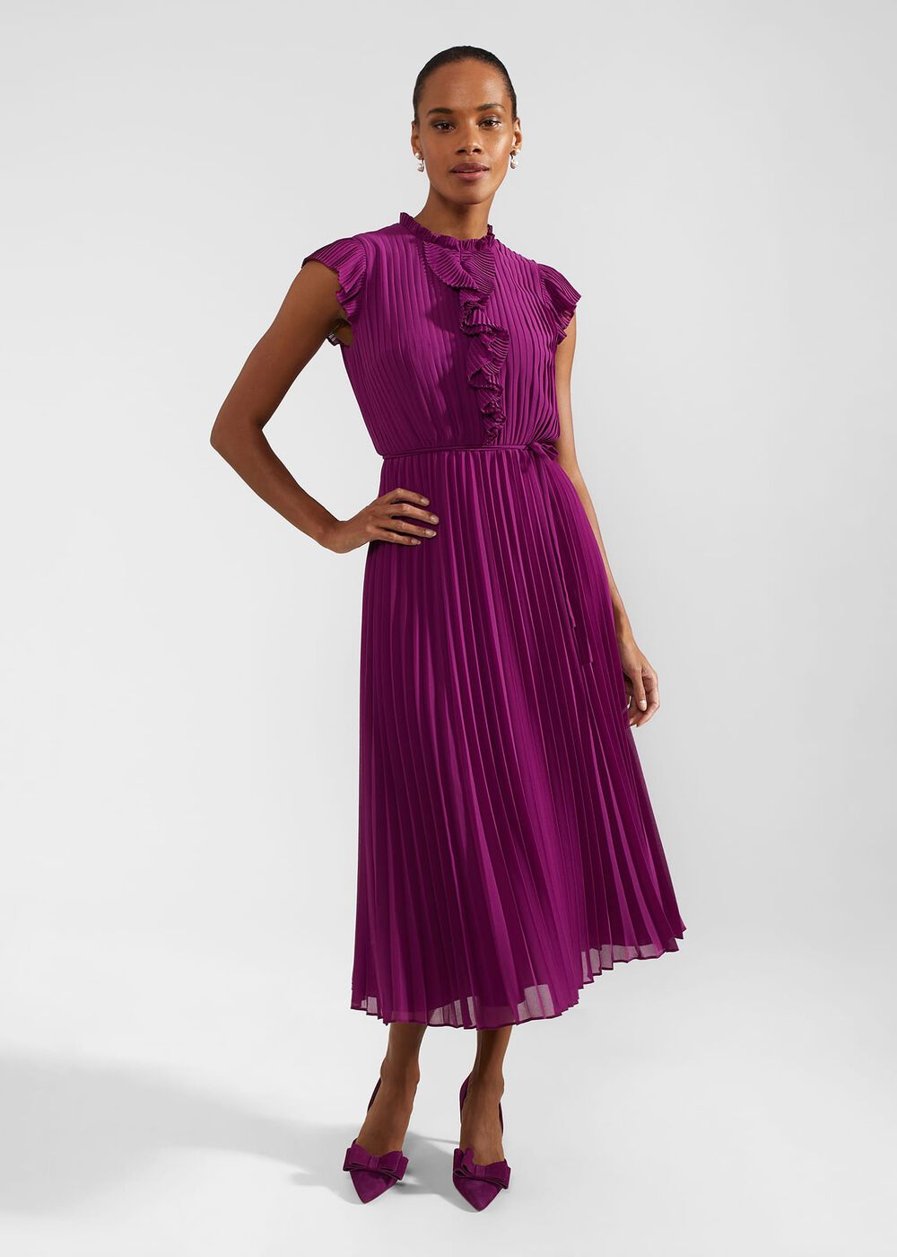 A woman wearing a purple pleated dress with a ruffled neckline and cap sleeves, standing against a plain background.