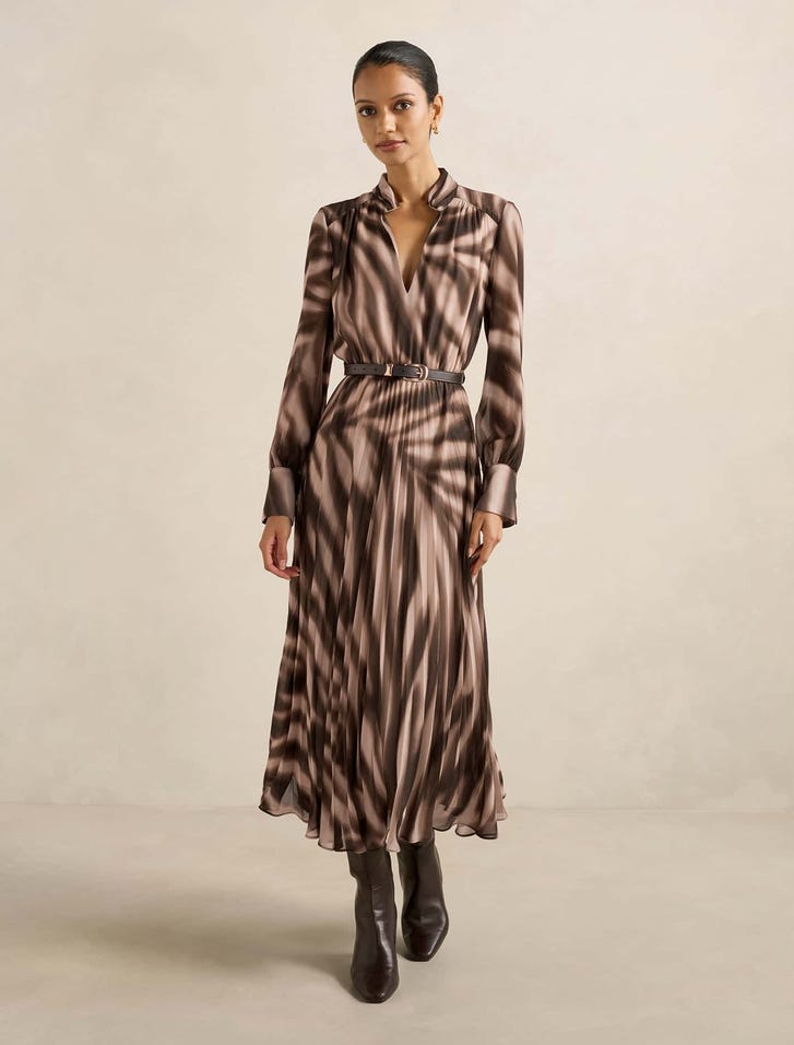 A woman wearing a brown and beige abstract print pleated midi dress with a stand collar, split neckline, long cuffs, and a waist belt, paired with dark boots.