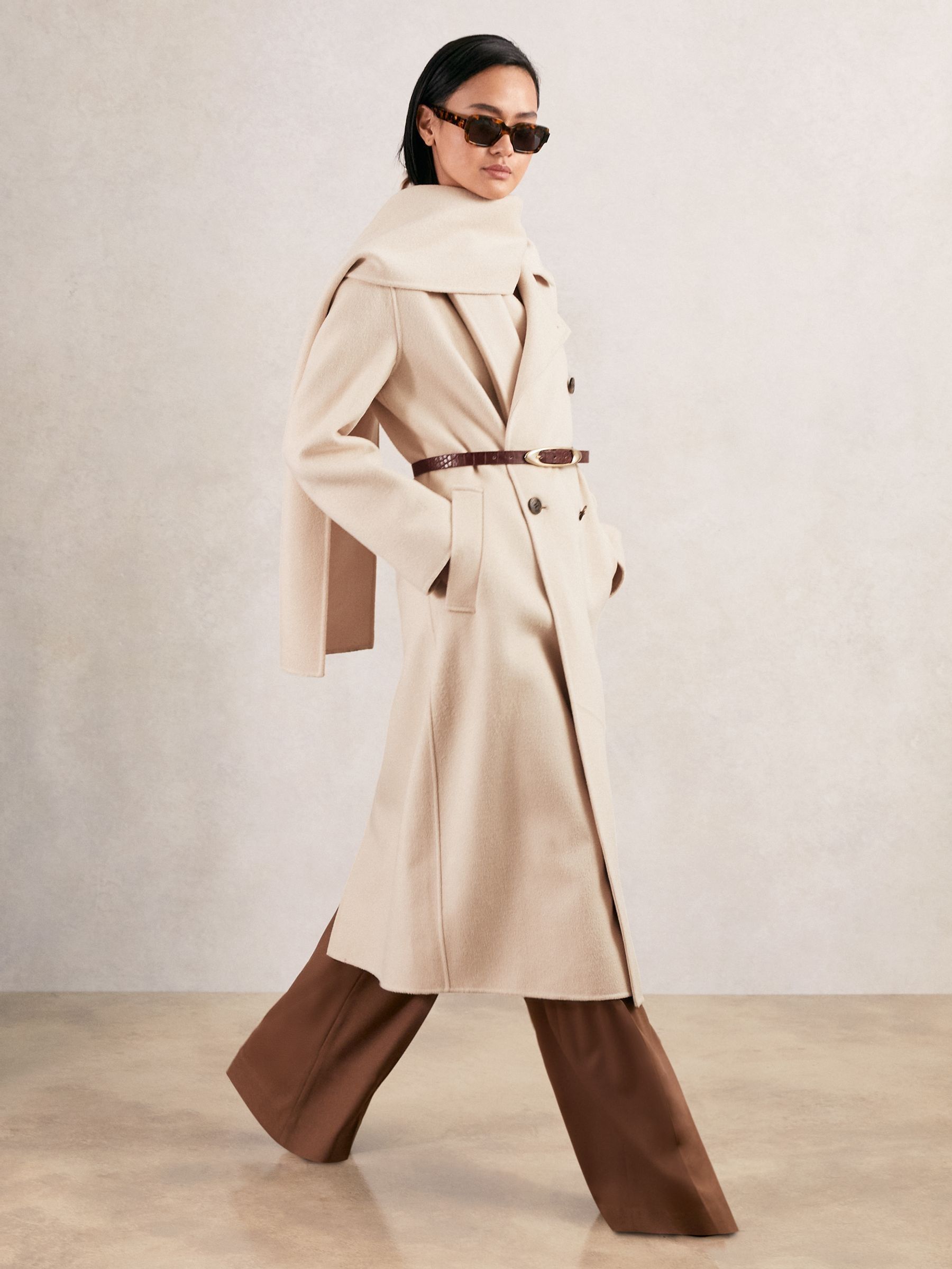A woman wearing a cream-colored, tailored wool-blend coat with a detachable scarf, six front buttons, and a petite fit, standing against a neutral background.