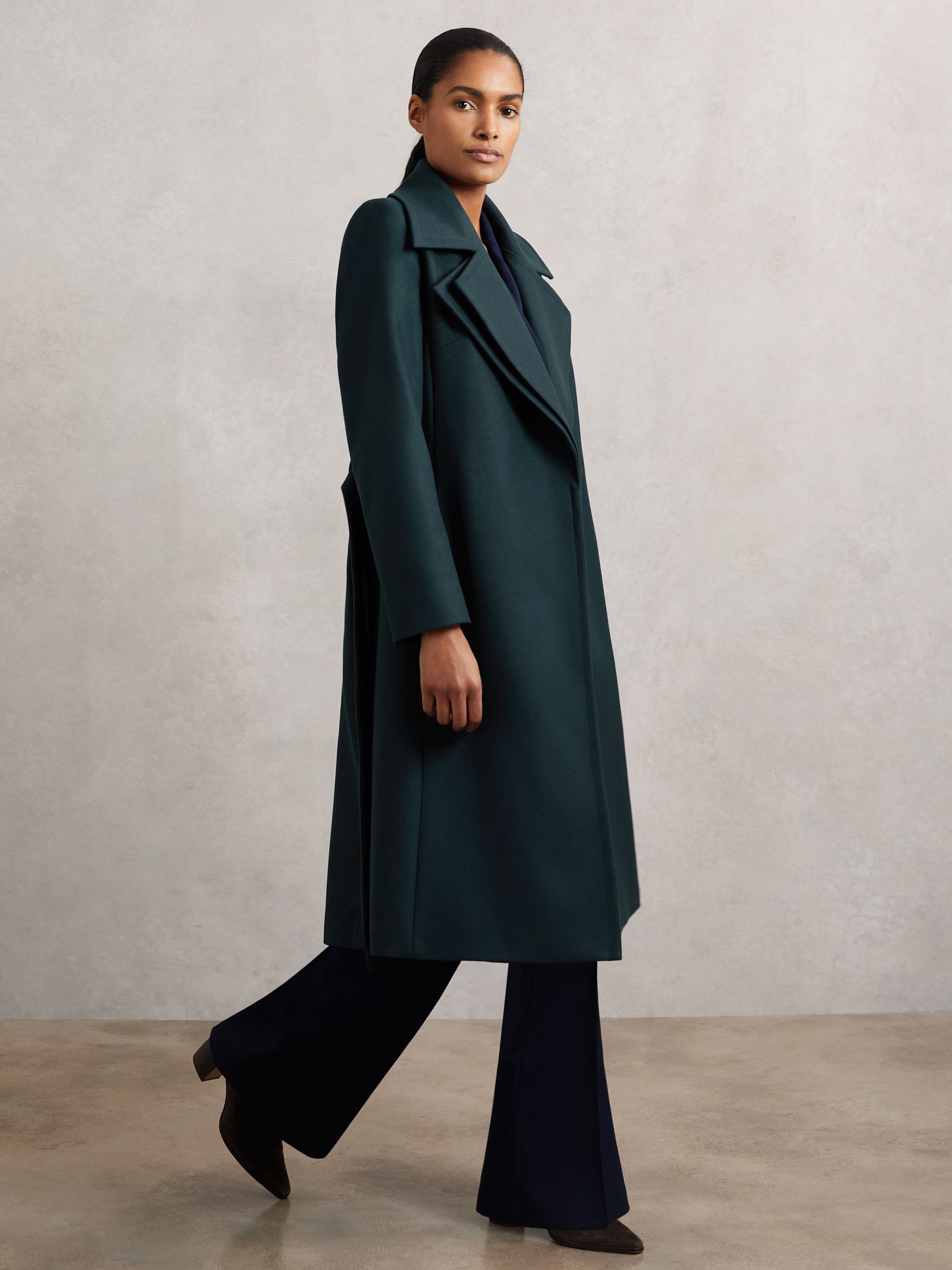 A woman wearing a teal wool-blend coat with double lapels, a belt, and side pockets, standing against a plain background.