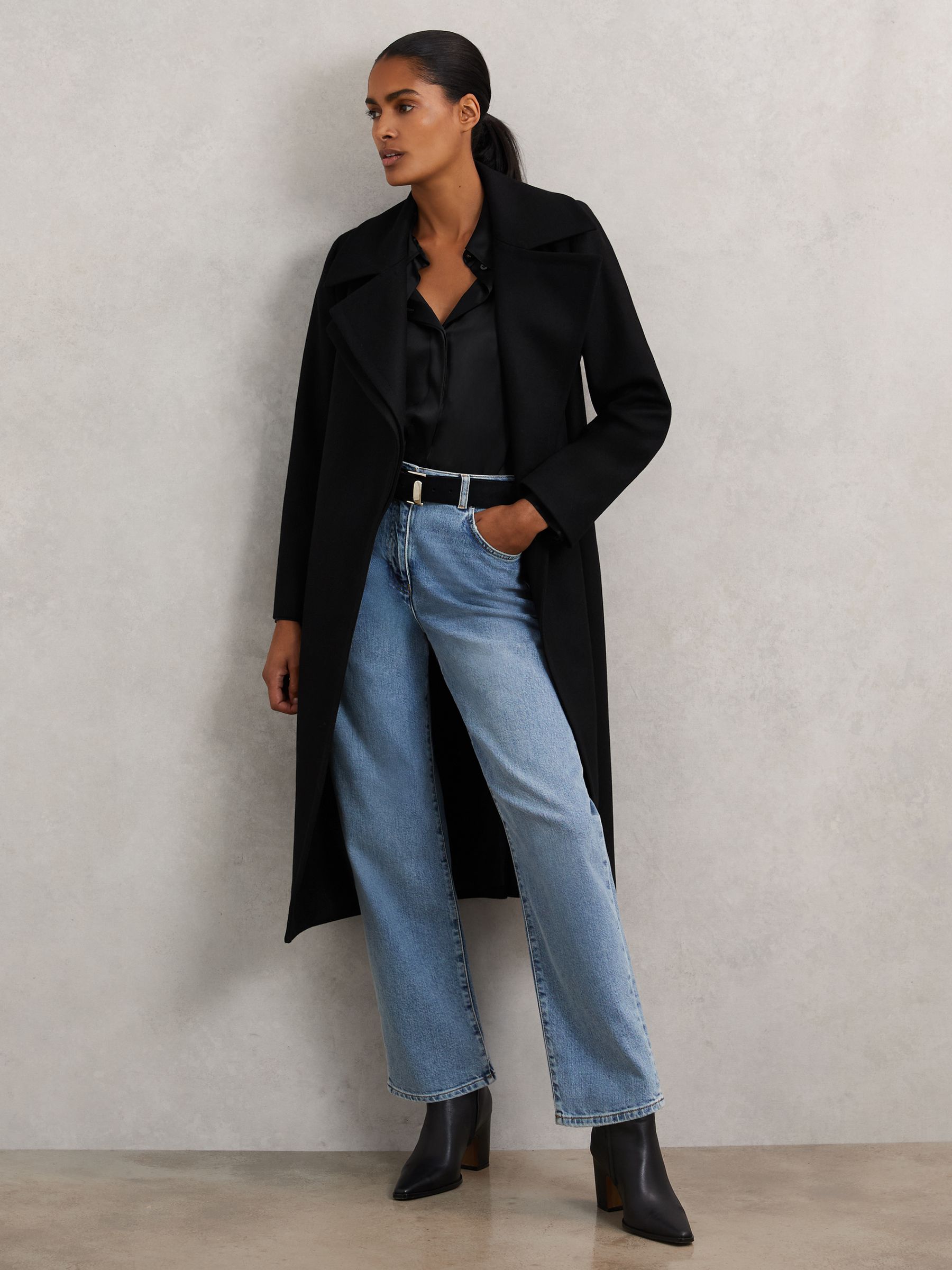A woman wearing a black wool-blend coat with double lapels, a belt, and side pockets, paired with jeans and black boots.
