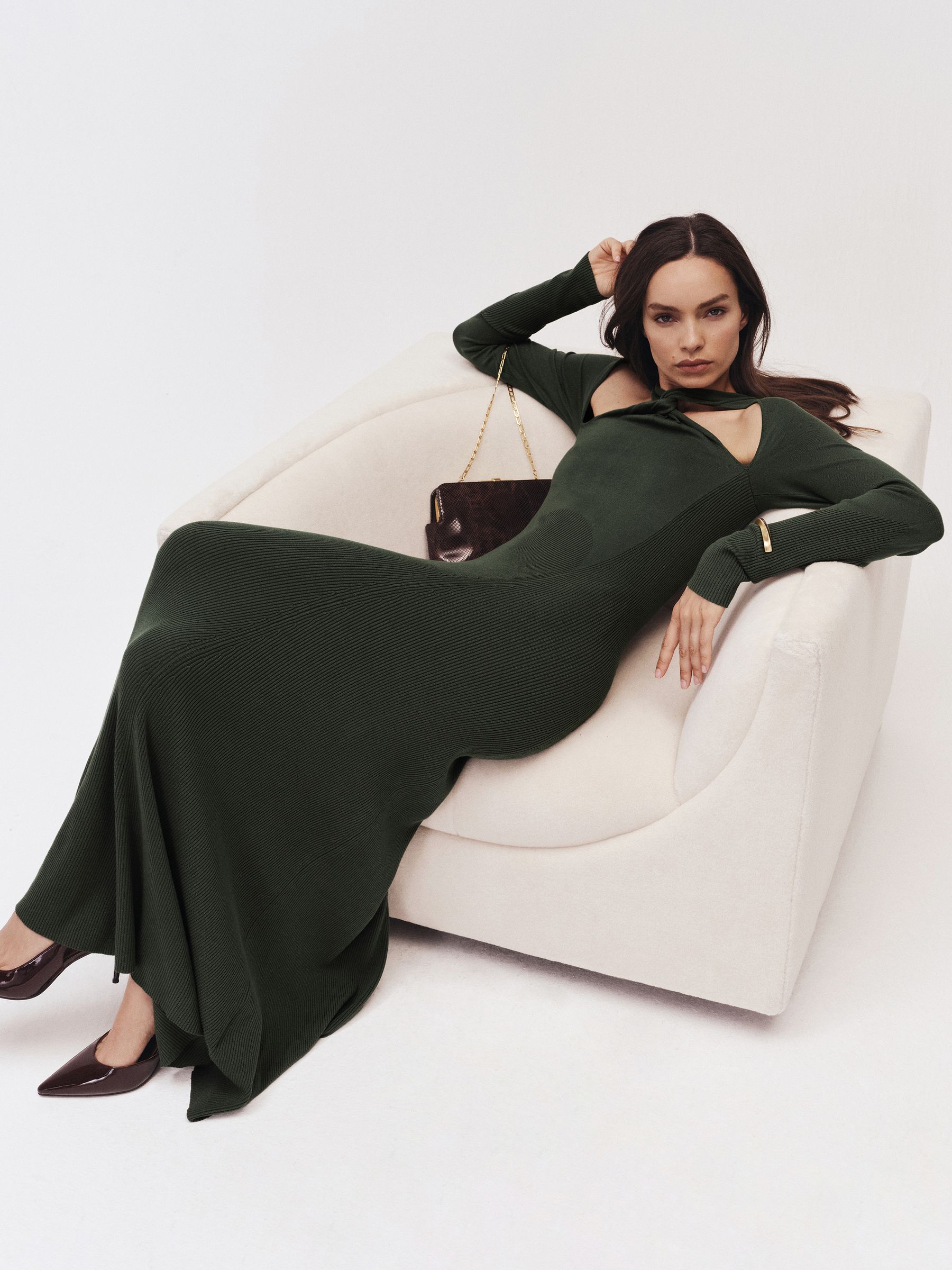 A woman lying on a white sofa wearing a dark green knitted maxi dress with cut-outs at the chest and back, long sleeves, and a high neckline.